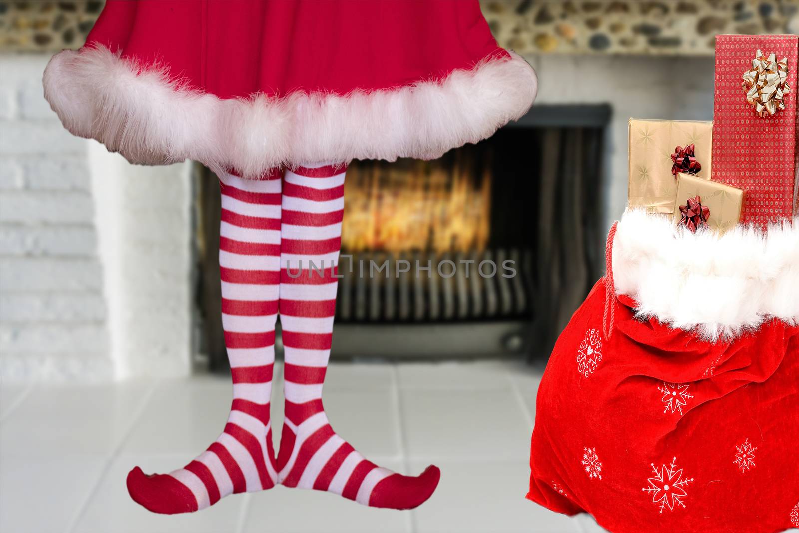 adorable cute little christmas elf girl with pointy feet wearing striped elf stockings and a red dress standing next to a santa claus bag full of presents in front of a burning fireplace