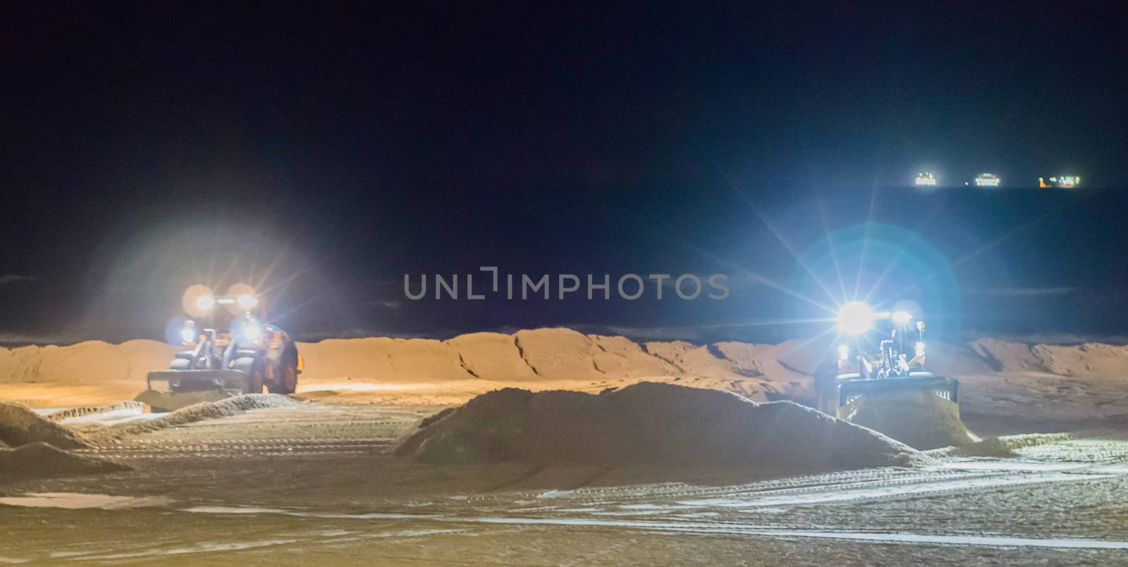 two ground workers working at night in bulldozers at the beach moving the sand