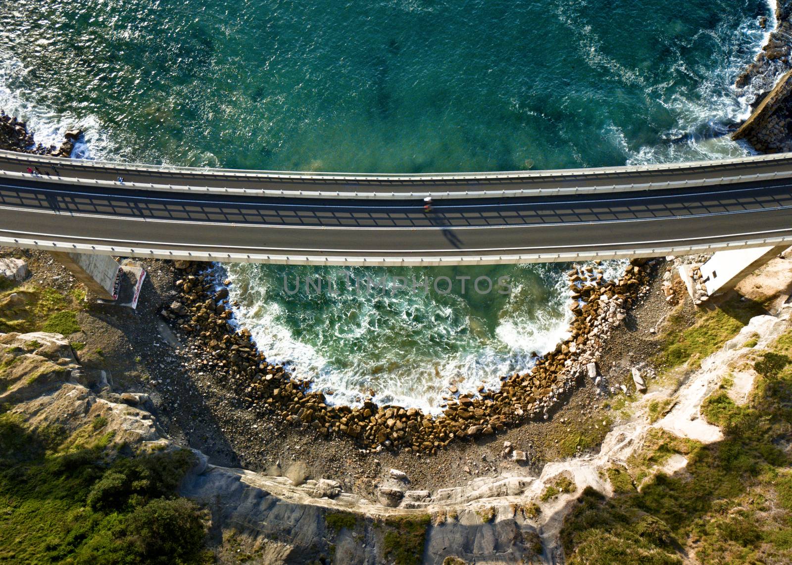 High above near the cliffs views down onto the Sea Cliff Bridge as a cyclist crosses leaving long shadows in the early morning light