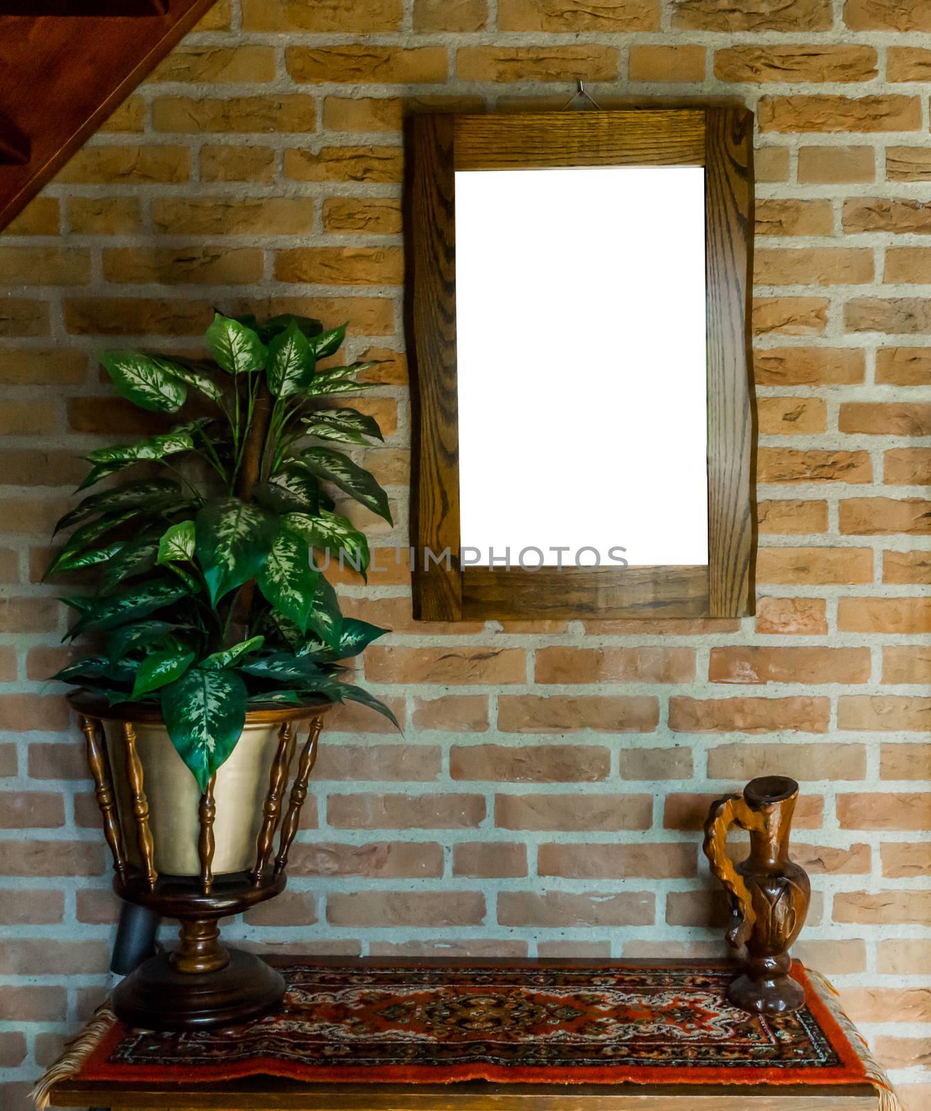 empty blank cut out wooden painting or mirror frame hanging on a brick wall above a table cabinet with a carpet rug decorated with a house plant and vase background texture