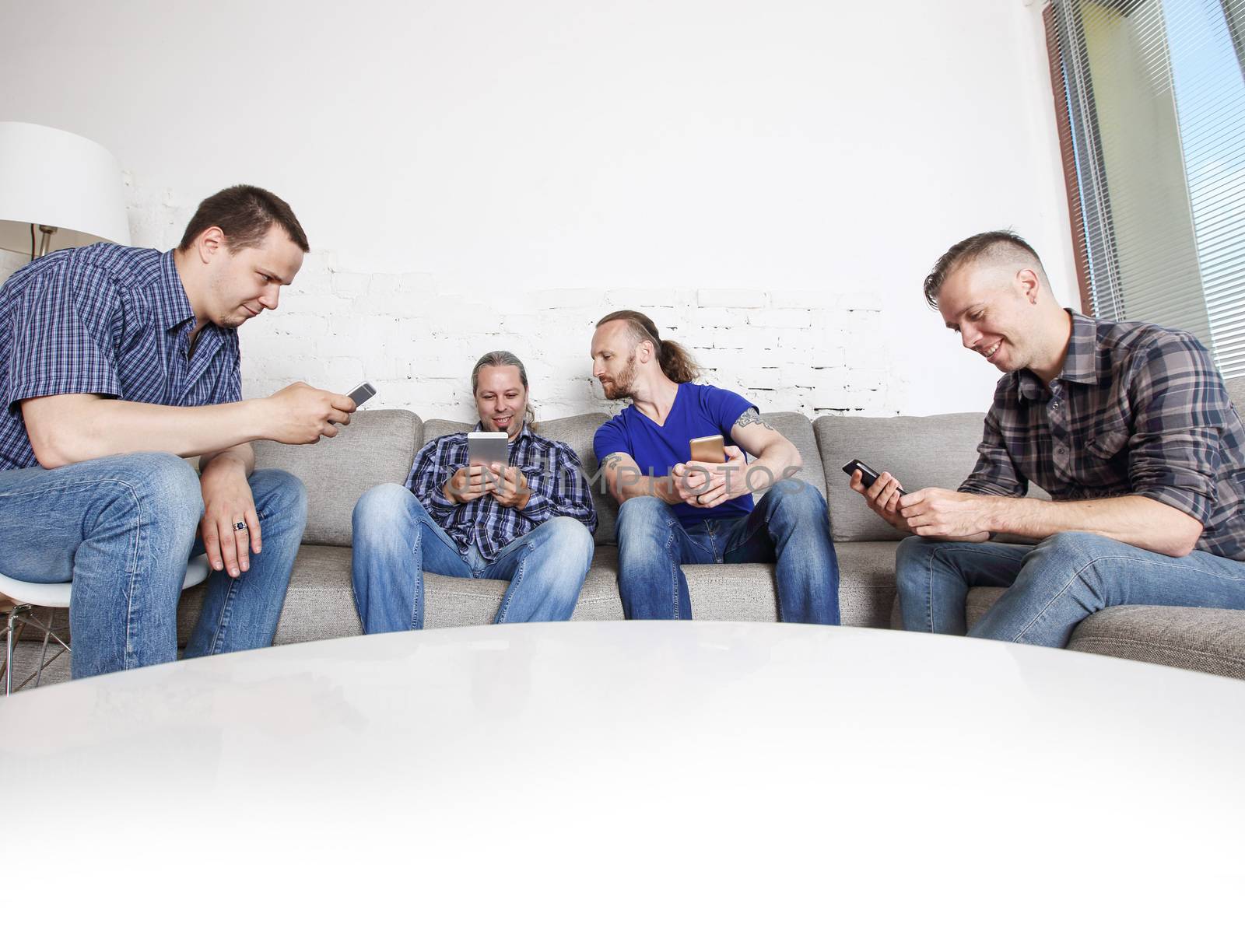 Four happy male friends using their mobile phones sitting on a sofa at home