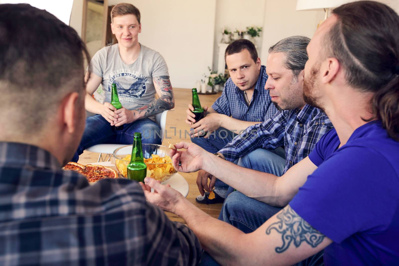 Group of men drinking beer, eating pizza, talking and smiling while resting at home on couch behind TV