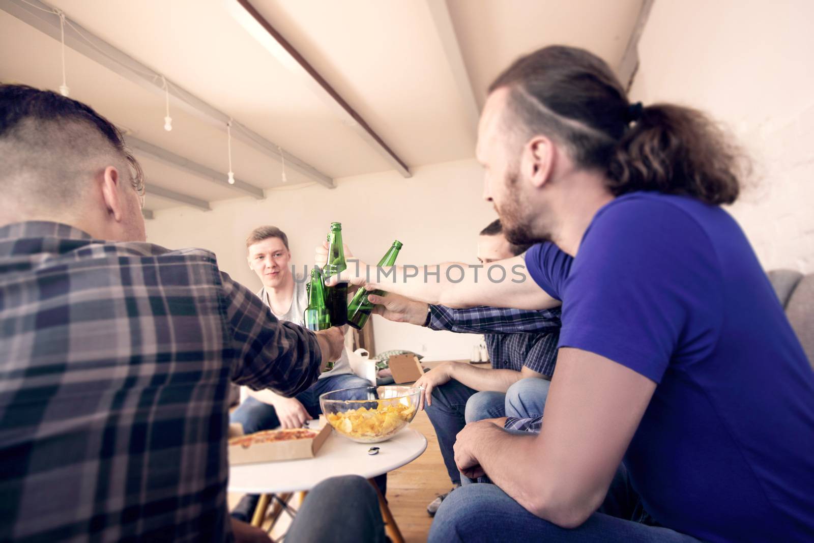 Group of men clinking beer, eating pizza, talking and smiling while resting at home on couch behind TV