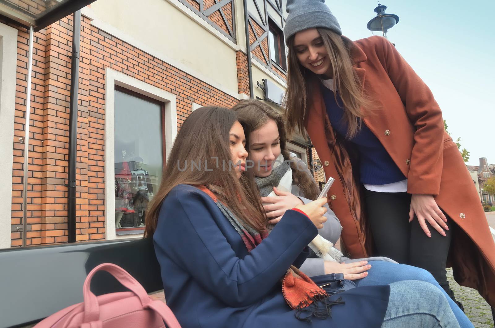 Three girlfriends sit on the bench and comment on the photo on the phone, smiling