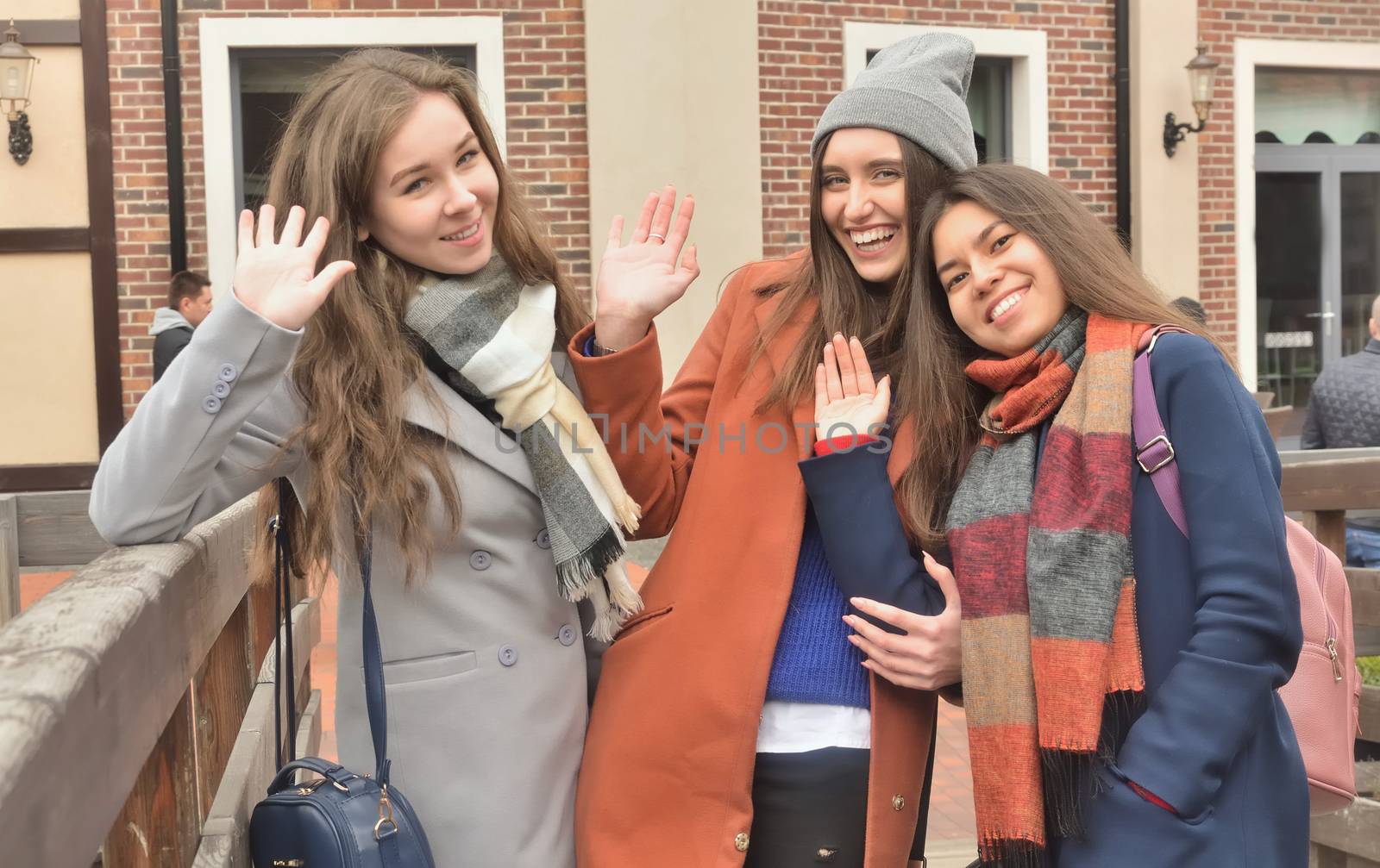 Three girlfriends in the coats stand together on the bridge, waving their hands and smiling at the camera