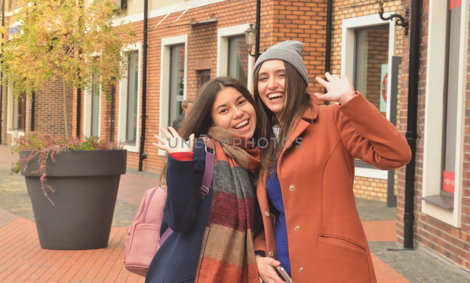 Two girlfriends in coats walking around the city, smiling and waving at the camera