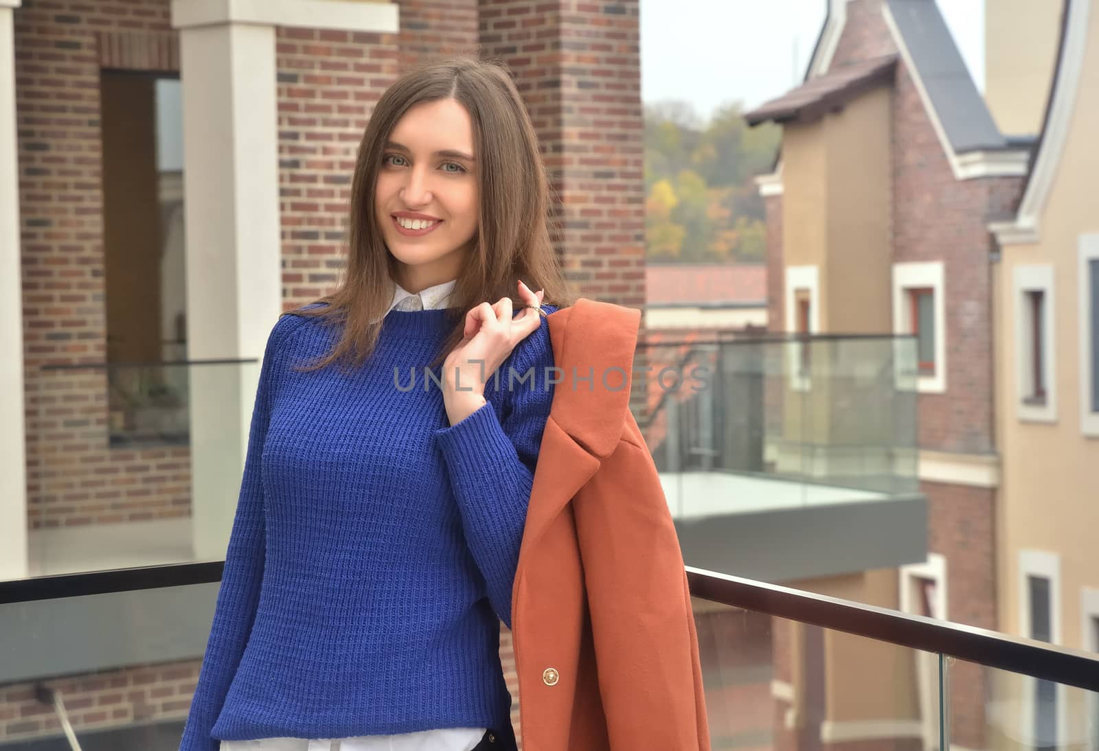 A beautiful girl standing against the backdrop of buildings, smiling at the camera and holding a coat in her hand