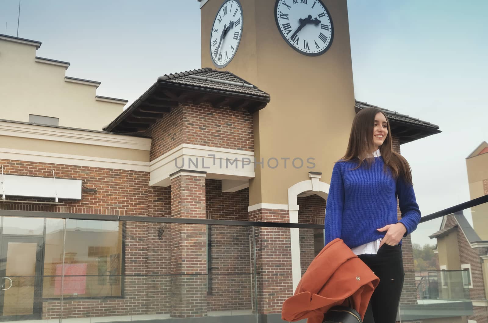 An attractive European girl stands on the balcony of a shopping center, against the backdrop of a street clock