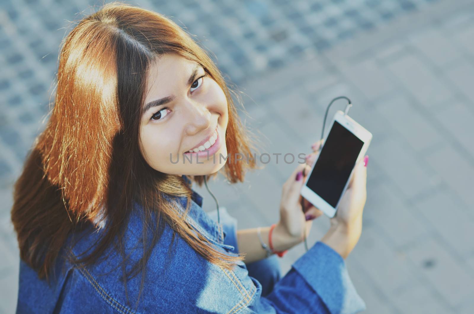 The student sits on a bench and listens to music on the phone with headphones, looks at the camera smiling, Asian appearance