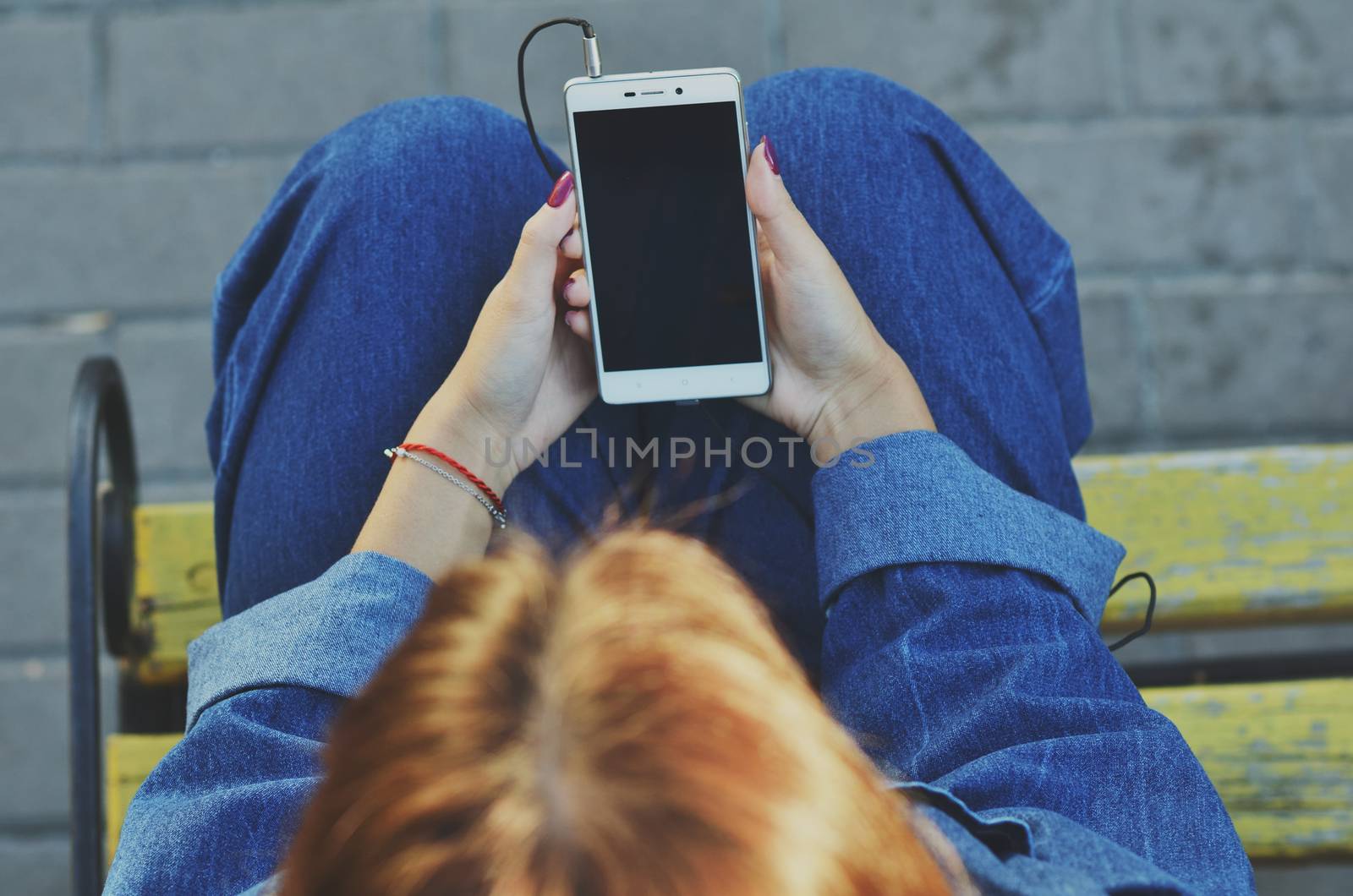 A young pretty student uses a smartphone sitting on a yellow bench in a denim jacket on the street