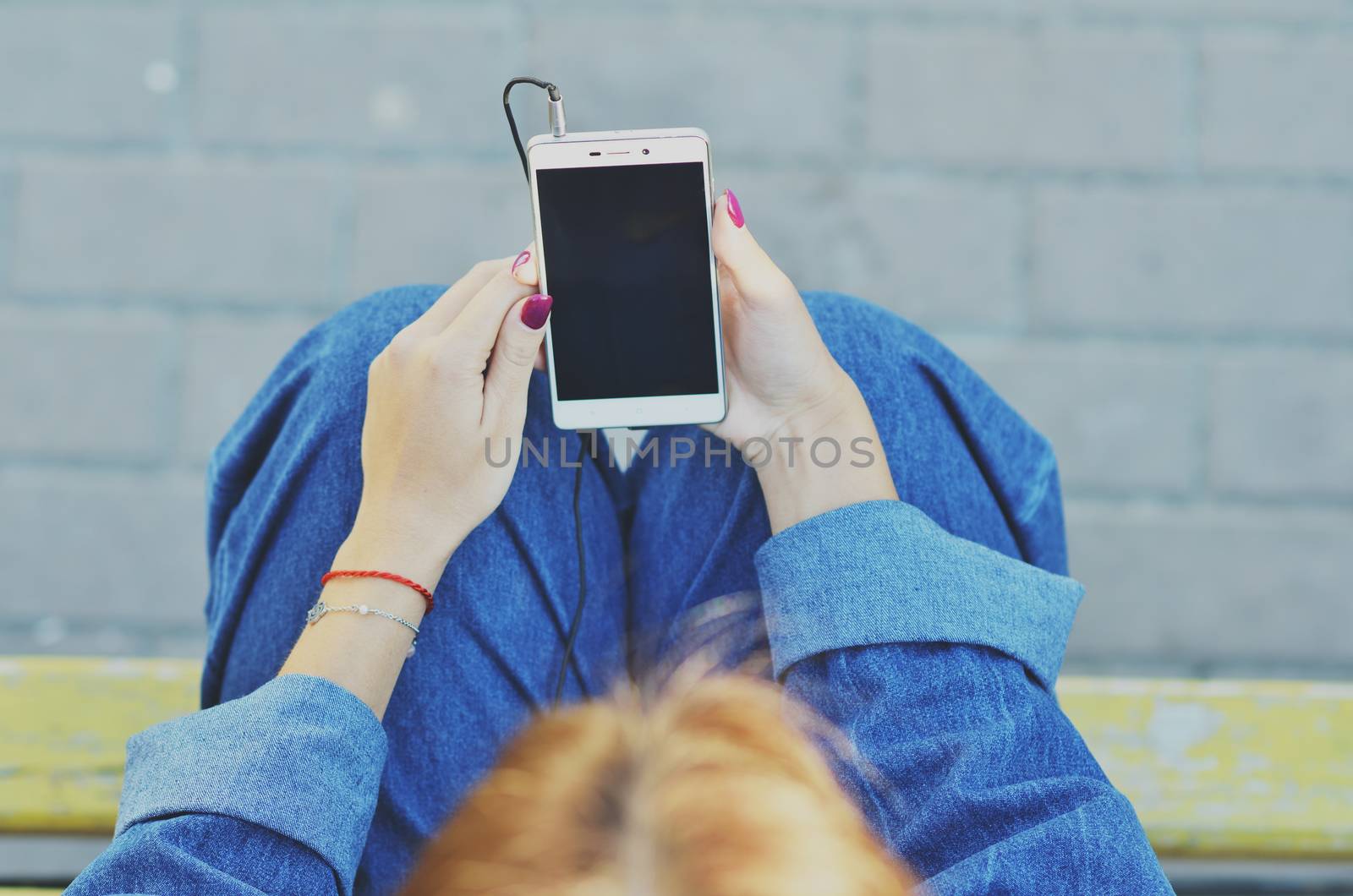 A young pretty student looks at the phone and listens to music in headphones sitting on a bench in the street