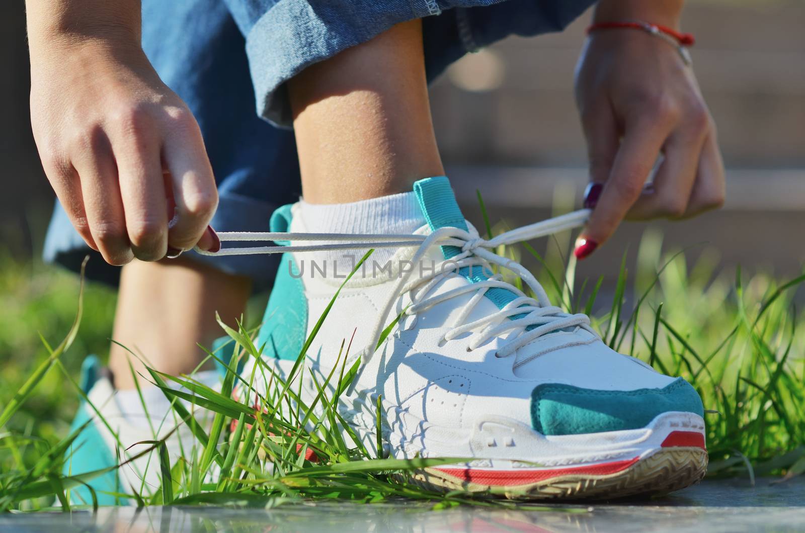 Young girl wearing jeans tying shoelaces on sneakers standing on green grass side view close-up horizontal photo, Sunny day