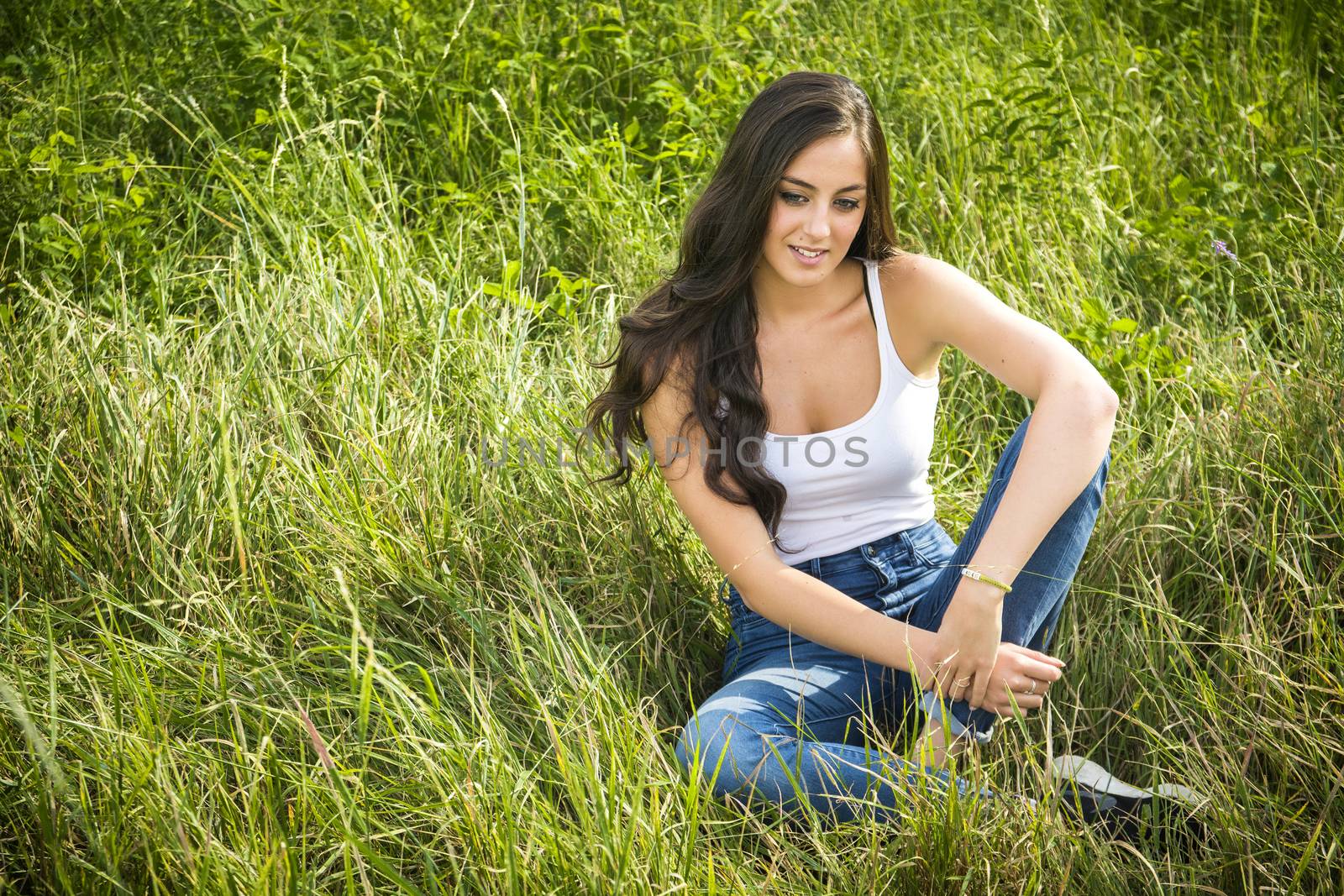 Portrait of beautiful smiling girl with long hair sitting on green grass and looking down