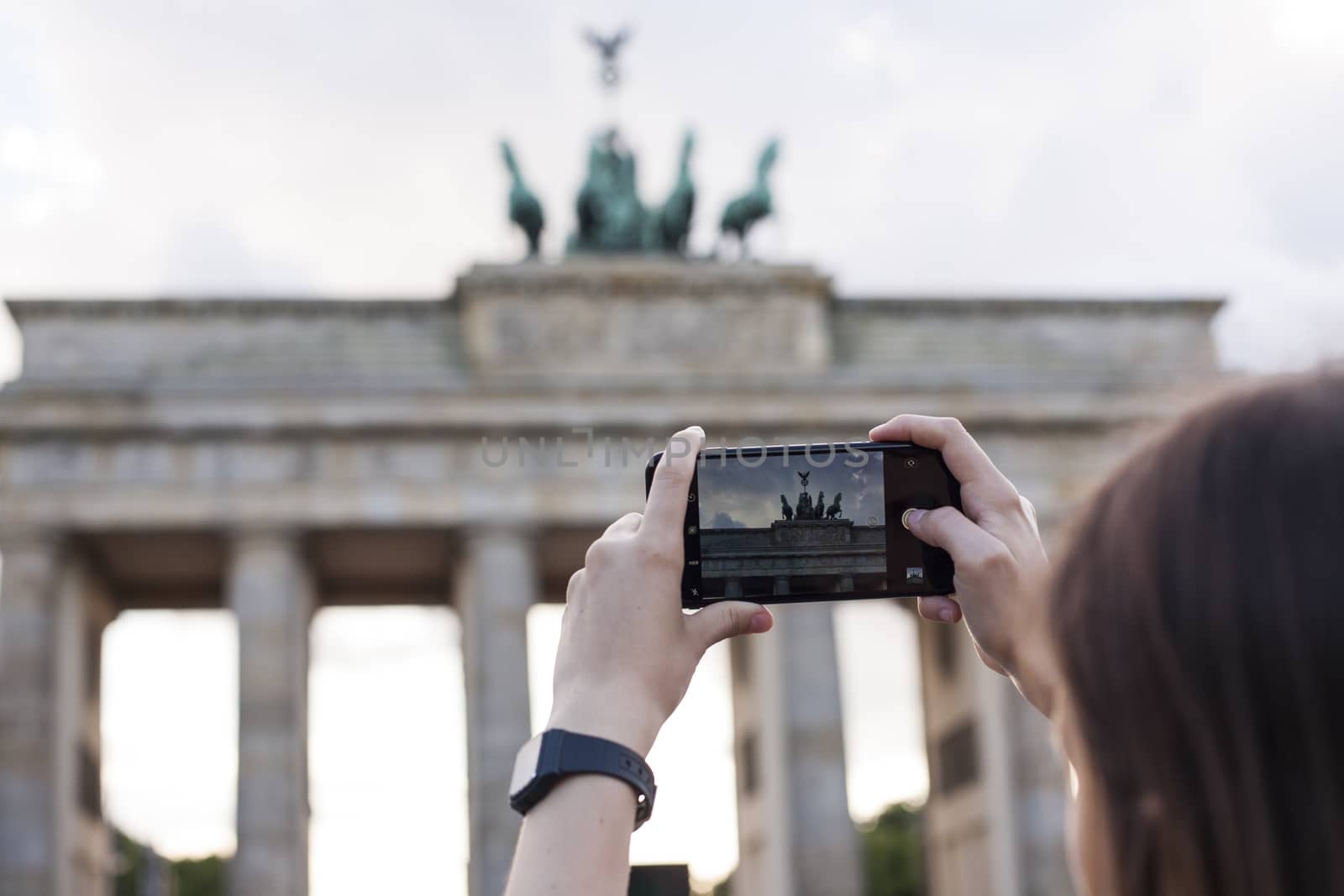 Hands holding phone mobile and takes pictures of famous historical tourist sites. The Brandenburg Gate neoclassical monument in Berlin.
