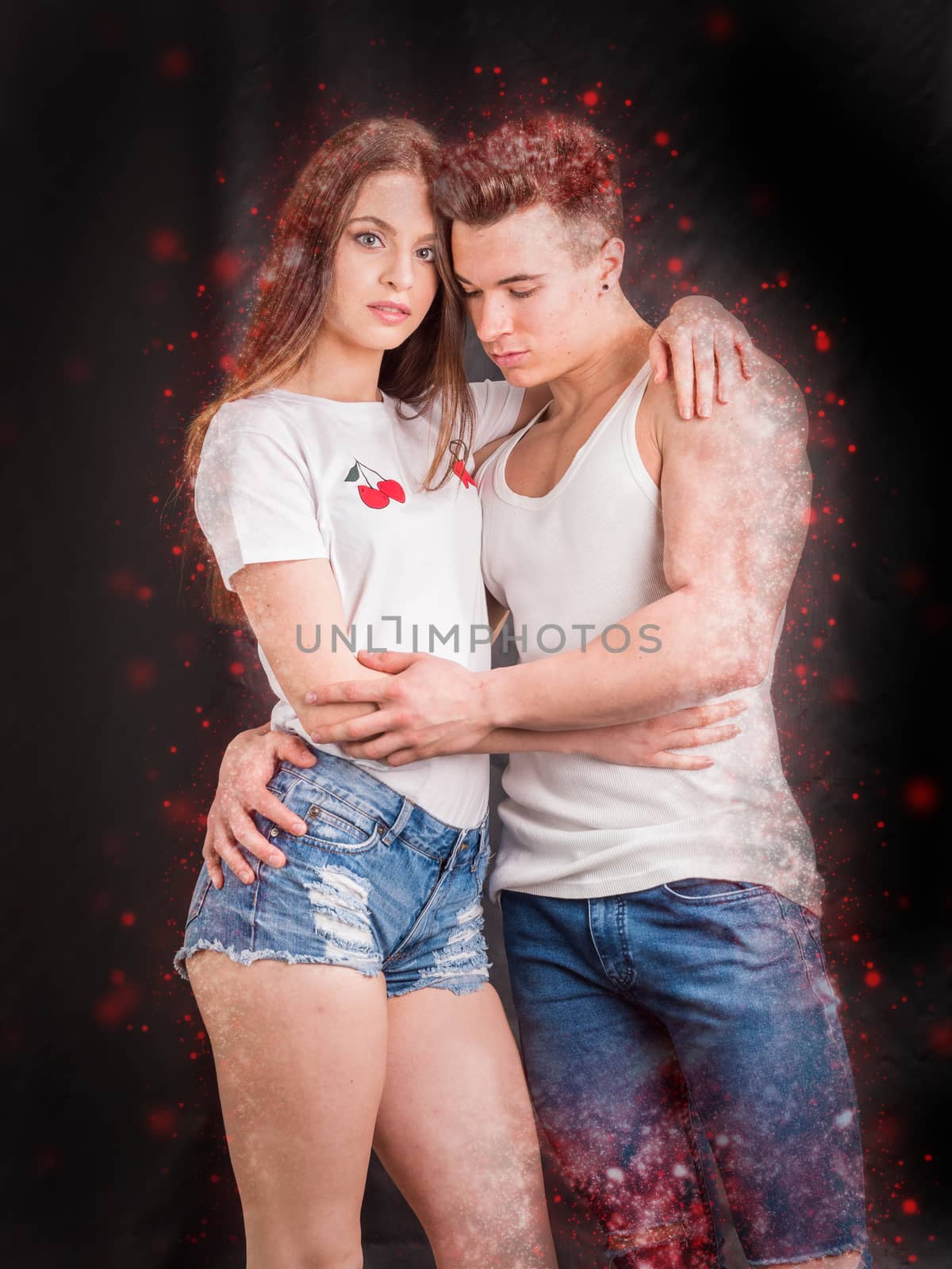 Romantic couple embracing in studio shot, man is muscular, woman is wearing denim shorts and white t-shirt, girl is looking at camera