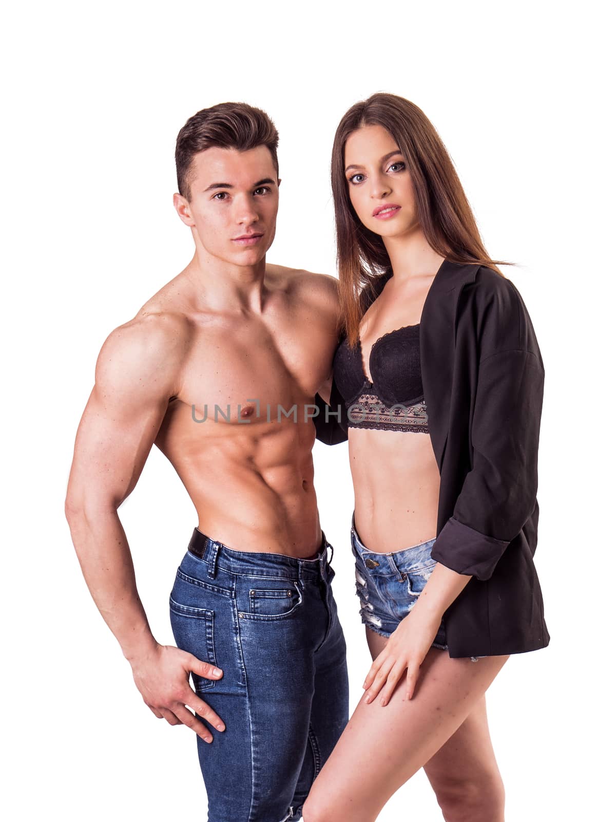 Romantic couple embracing in studio shot, man is muscular and shirtless, woman is showing bra under open shirt, both looking at camera