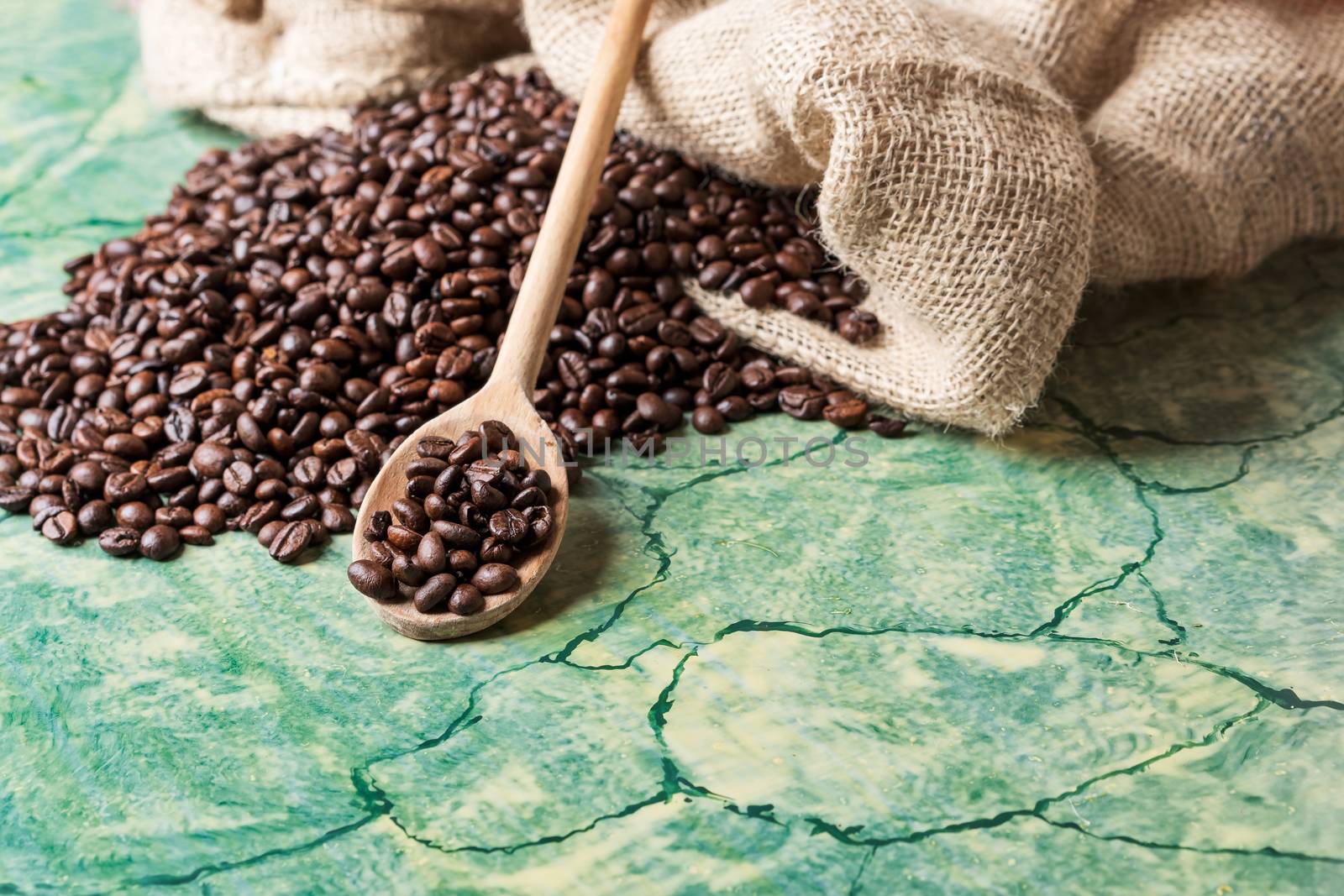 Coffee beans in coffee burlap bag on green table and wooden spoon with coffee beans on top.