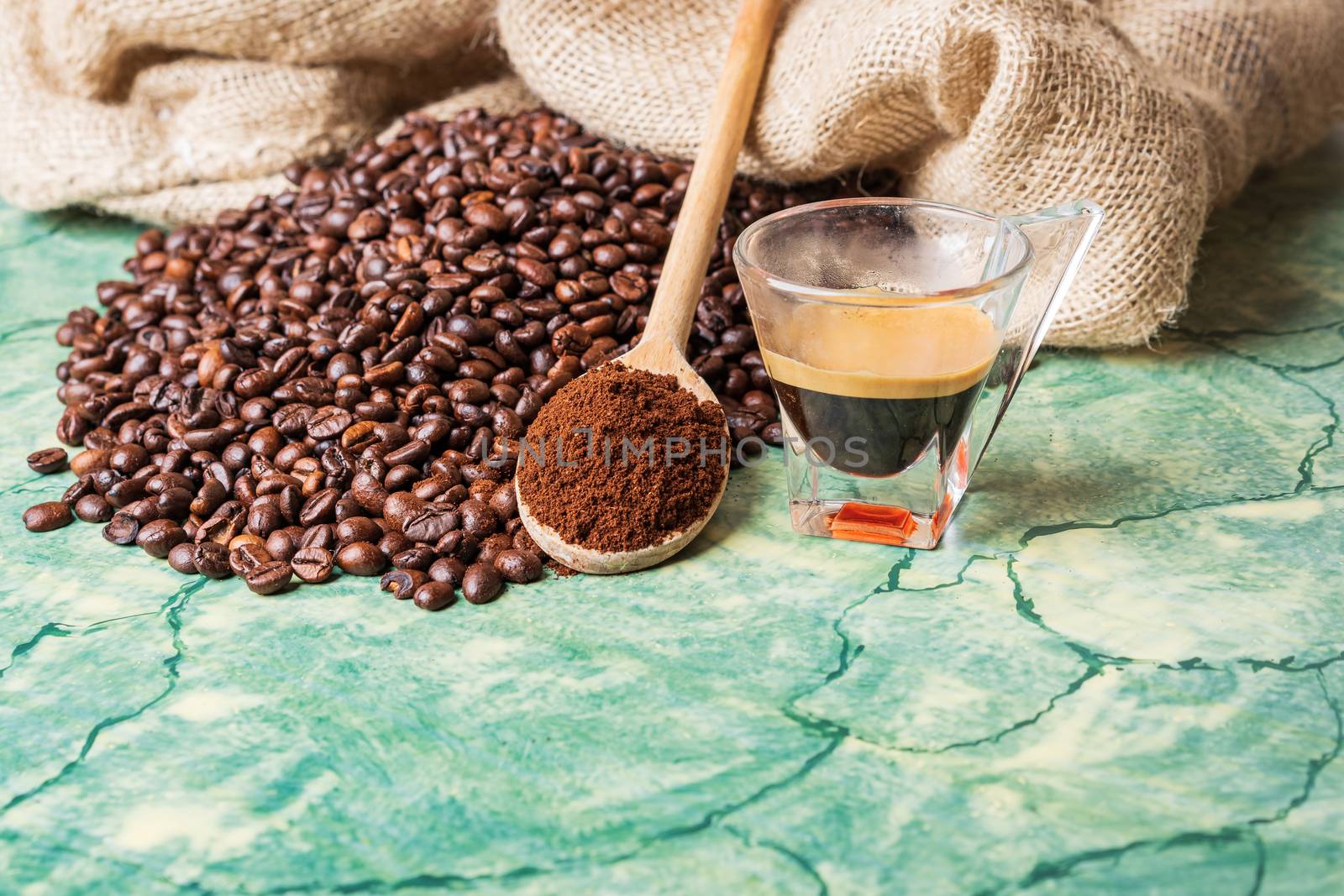 Coffee beans in coffee burlap bag on green surface,wooden spoon with ground coffee on top and coffee glass cup.