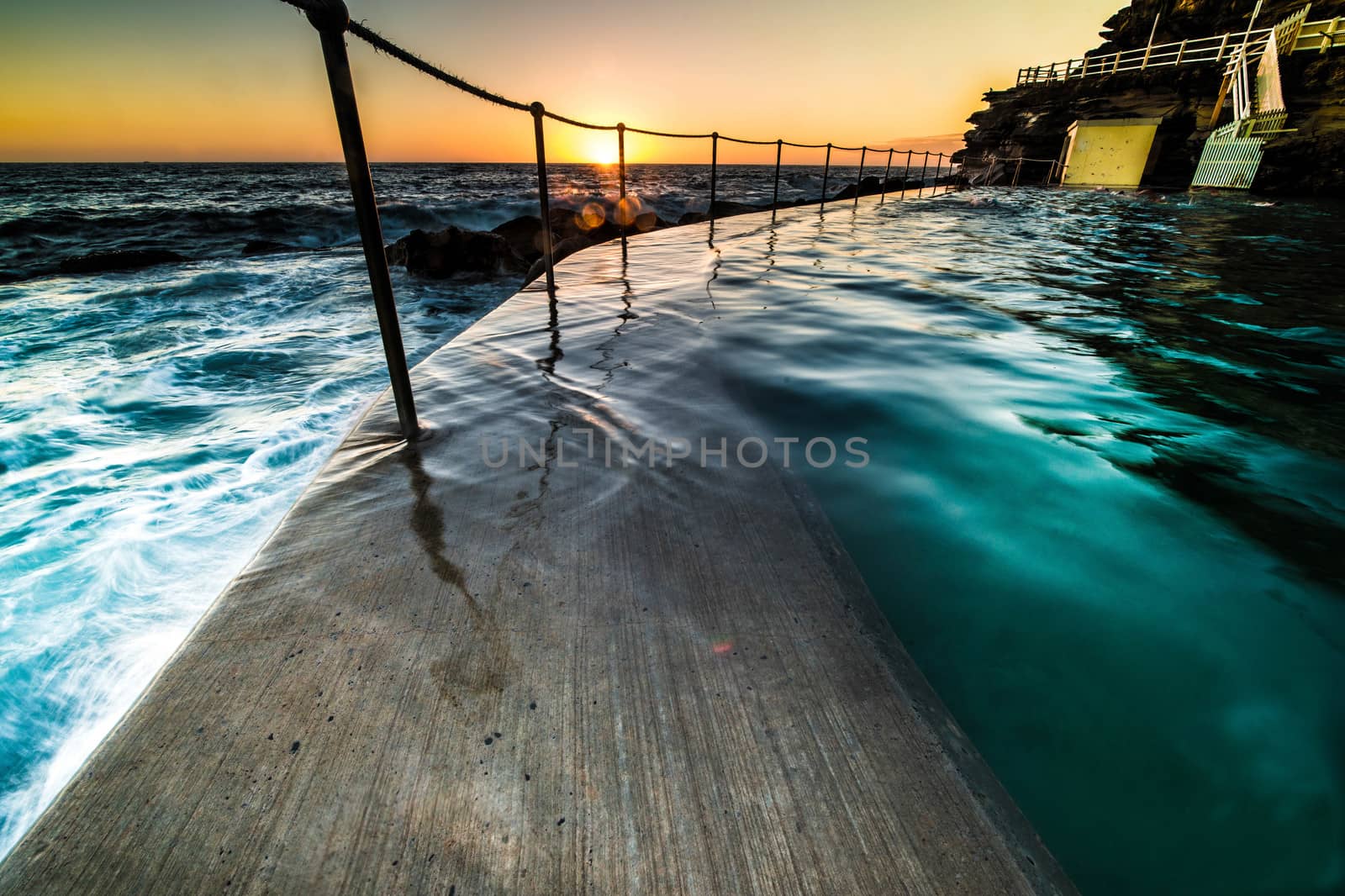 Bronte Pool, Sydney at sunrise with the orb of the sun just appearing over the horizon lighting up the rock pool as it juts out into the ocean with surf cascading over the wall
