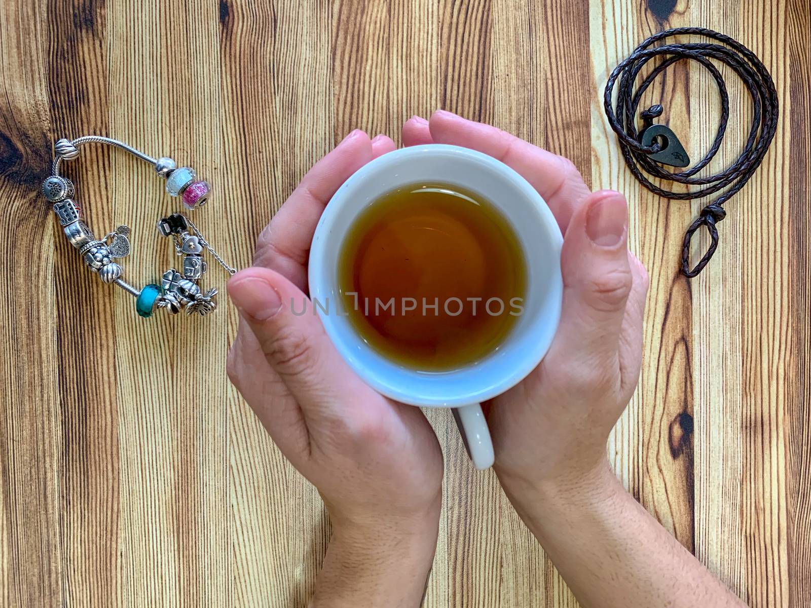 A closeup of a woman's hands holding a cup of hot tea. The background is wooden table. One of the bracelets is made of leather and the other is silver