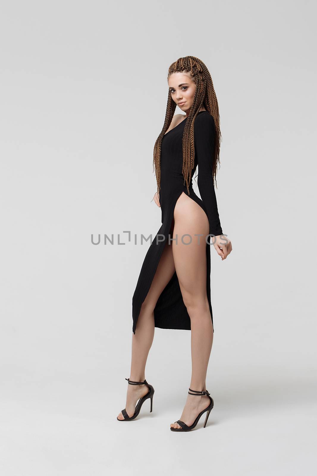 Side view of sensual young female with long dreadlocks wearing amazing black dress and looking at camera while standing on light gray background