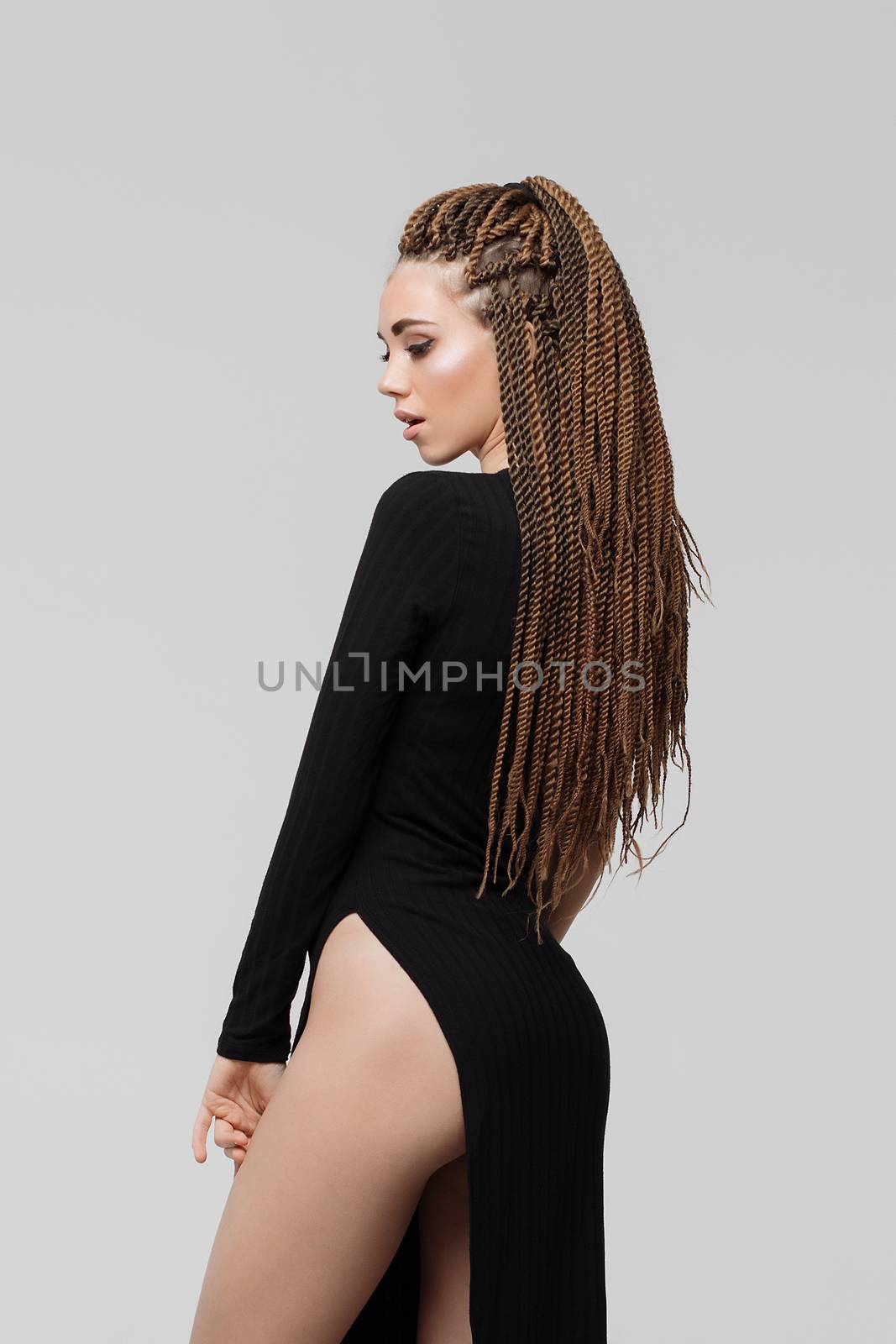 Side view of sensual young female with long dreadlocks wearing amazing black dress and looking at camera while standing on light gray background