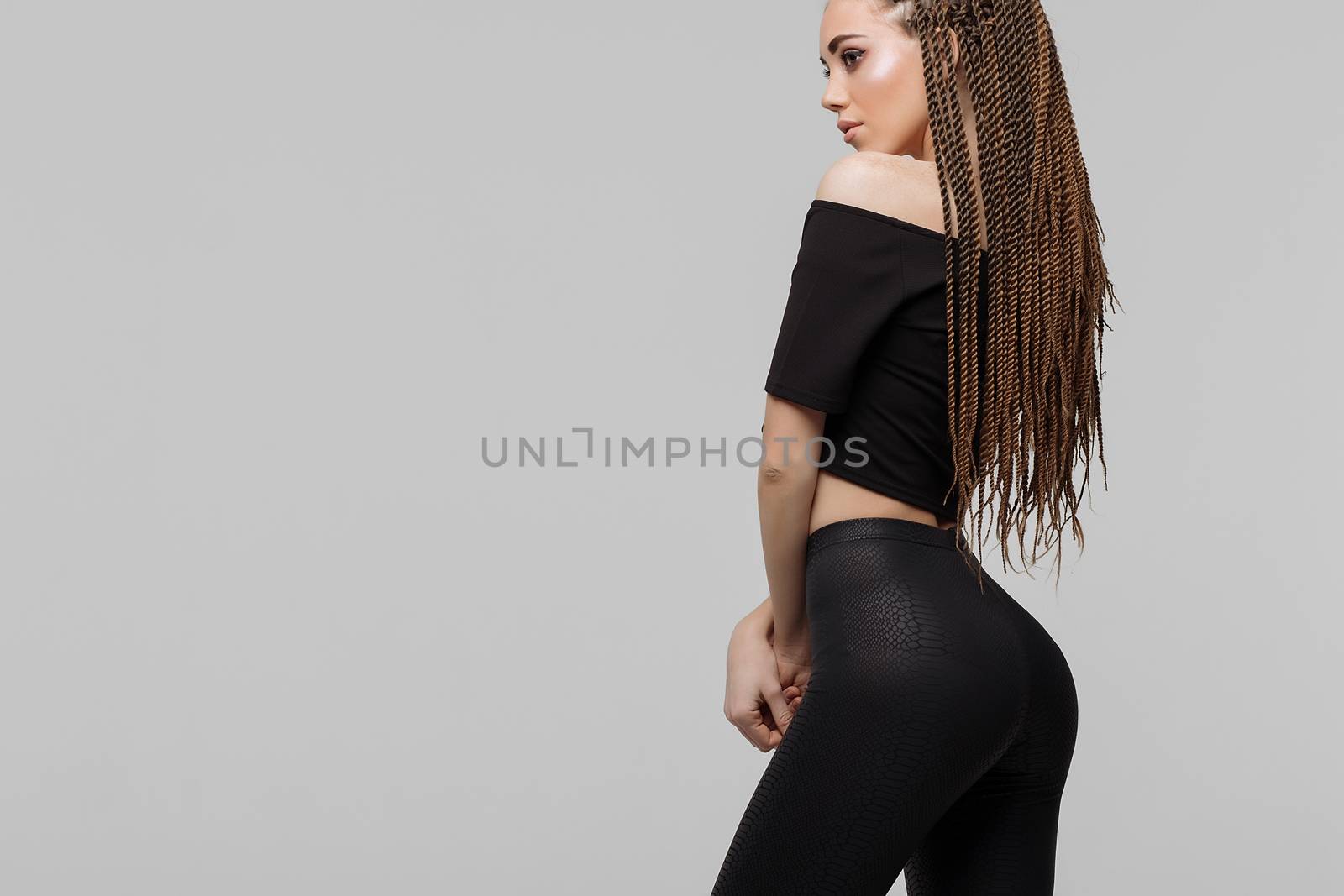 Beautiful young woman with dreadlocks keeping hand on waist and looking at camera while standing on light gray background