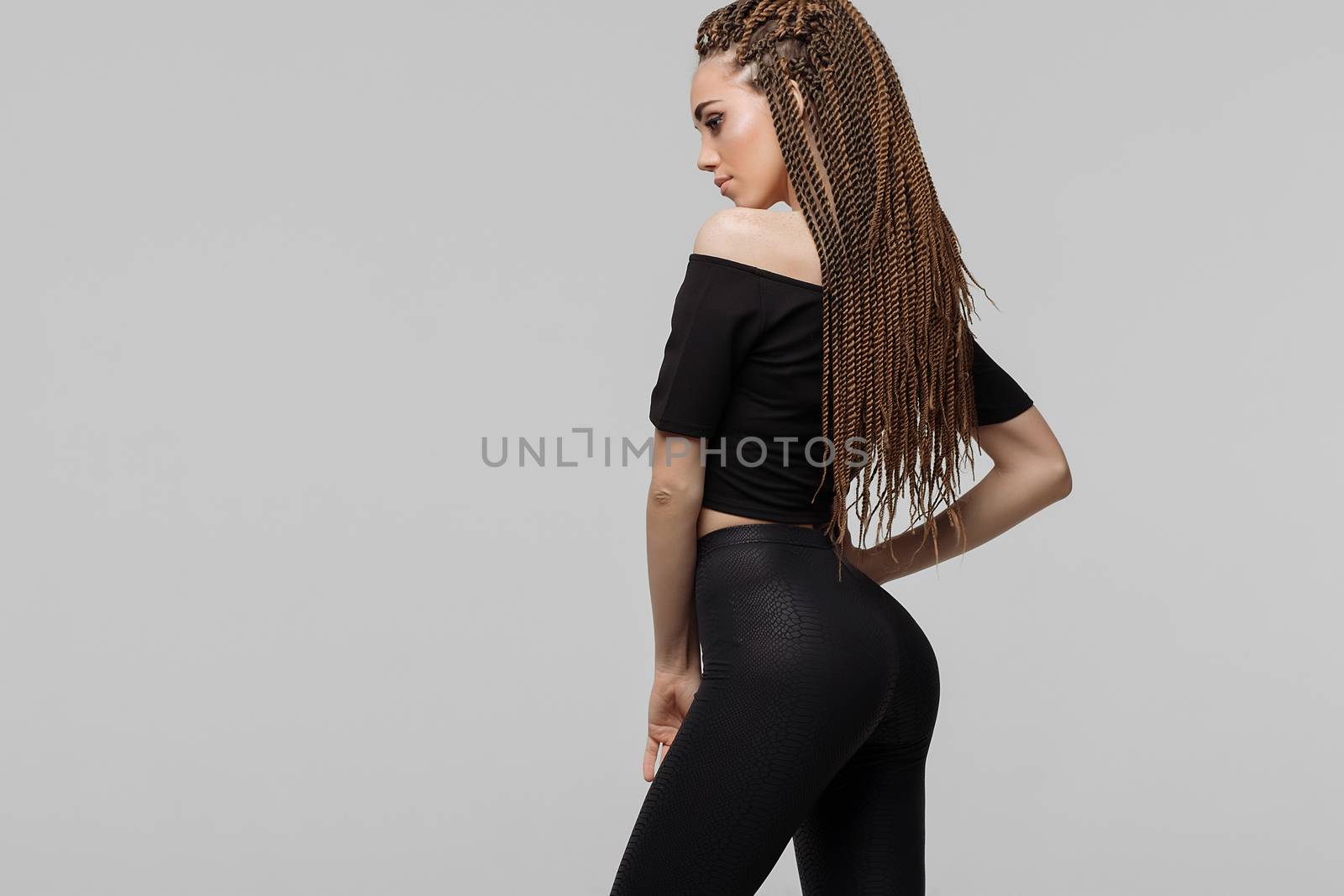 Beautiful young woman with dreadlocks keeping hand on waist and looking at camera while standing on light gray background