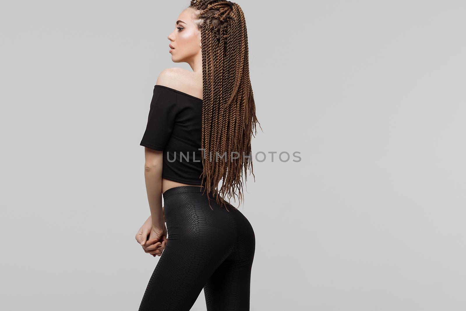 Beautiful young woman with dreadlocks keeping hand on waist and looking at camera while standing on light gray background