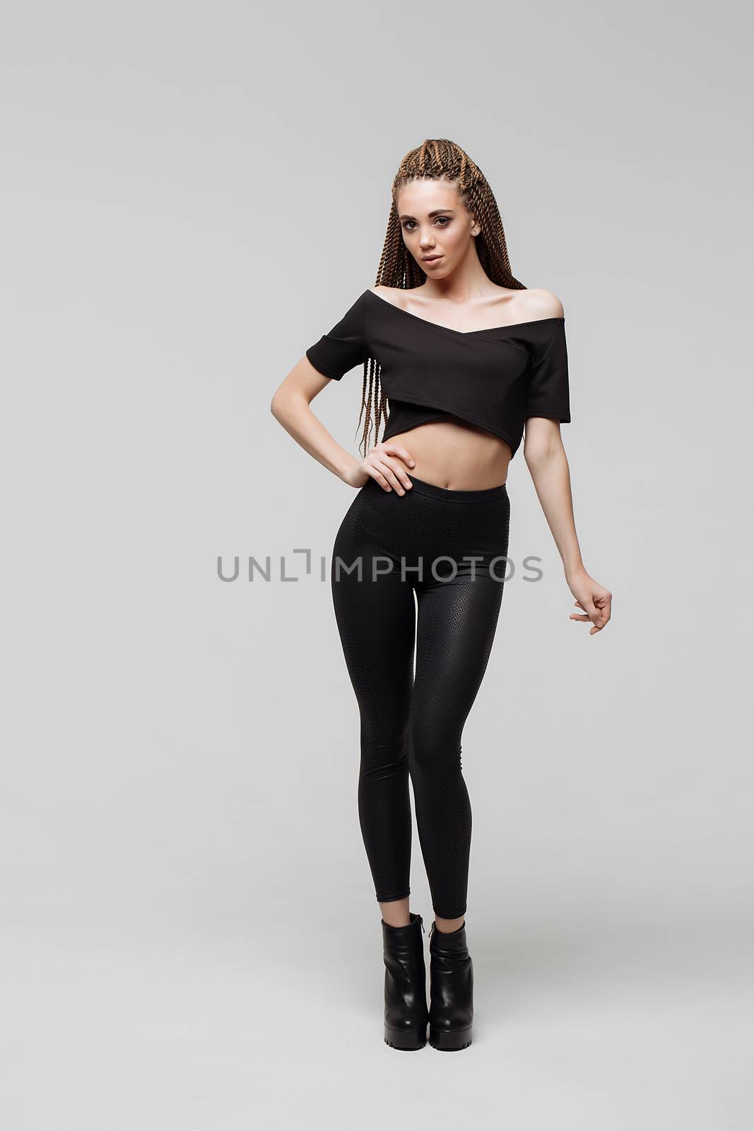Beautiful young woman with dreadlocks keeping hand on waist and looking at camera while standing on light gray background