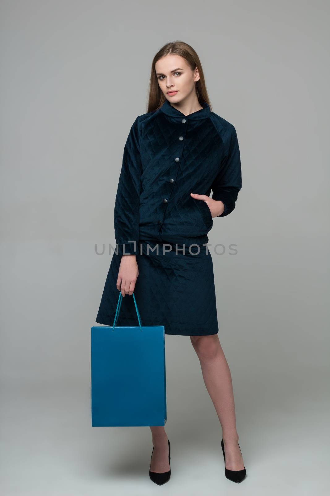 Young model long-haired blond girl in dark skirt suit stands holding blue shopping package