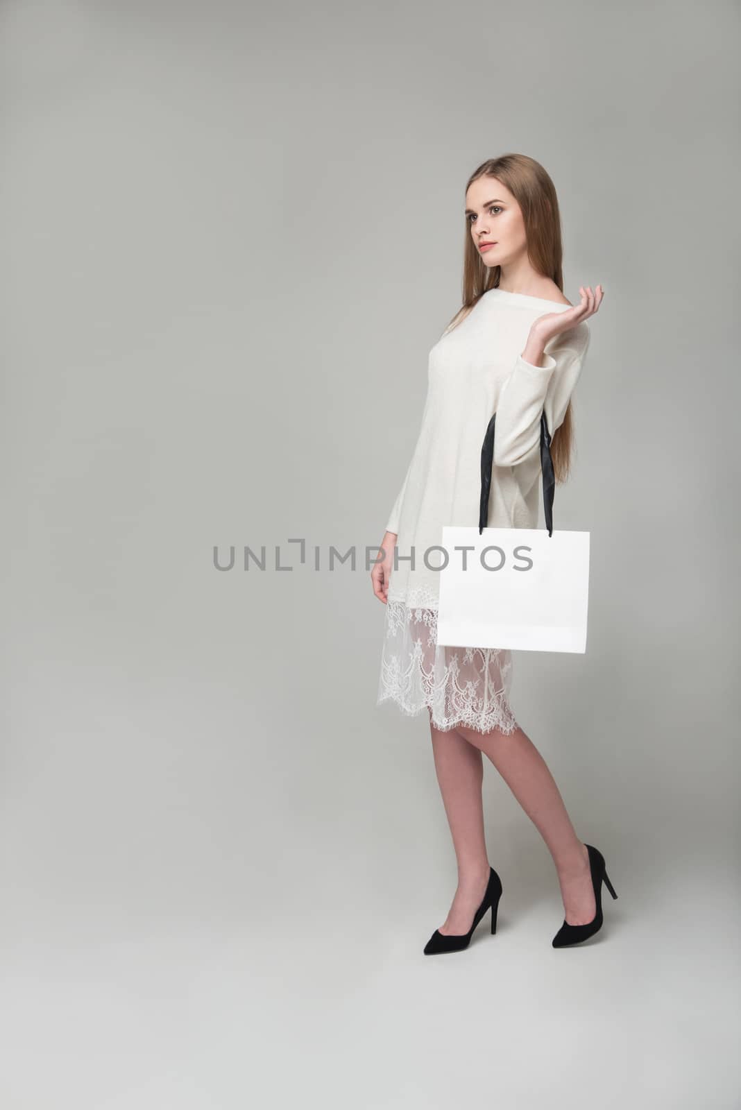 Young model long-haired blond girl in white short dress with lace stands holding white package with copy space