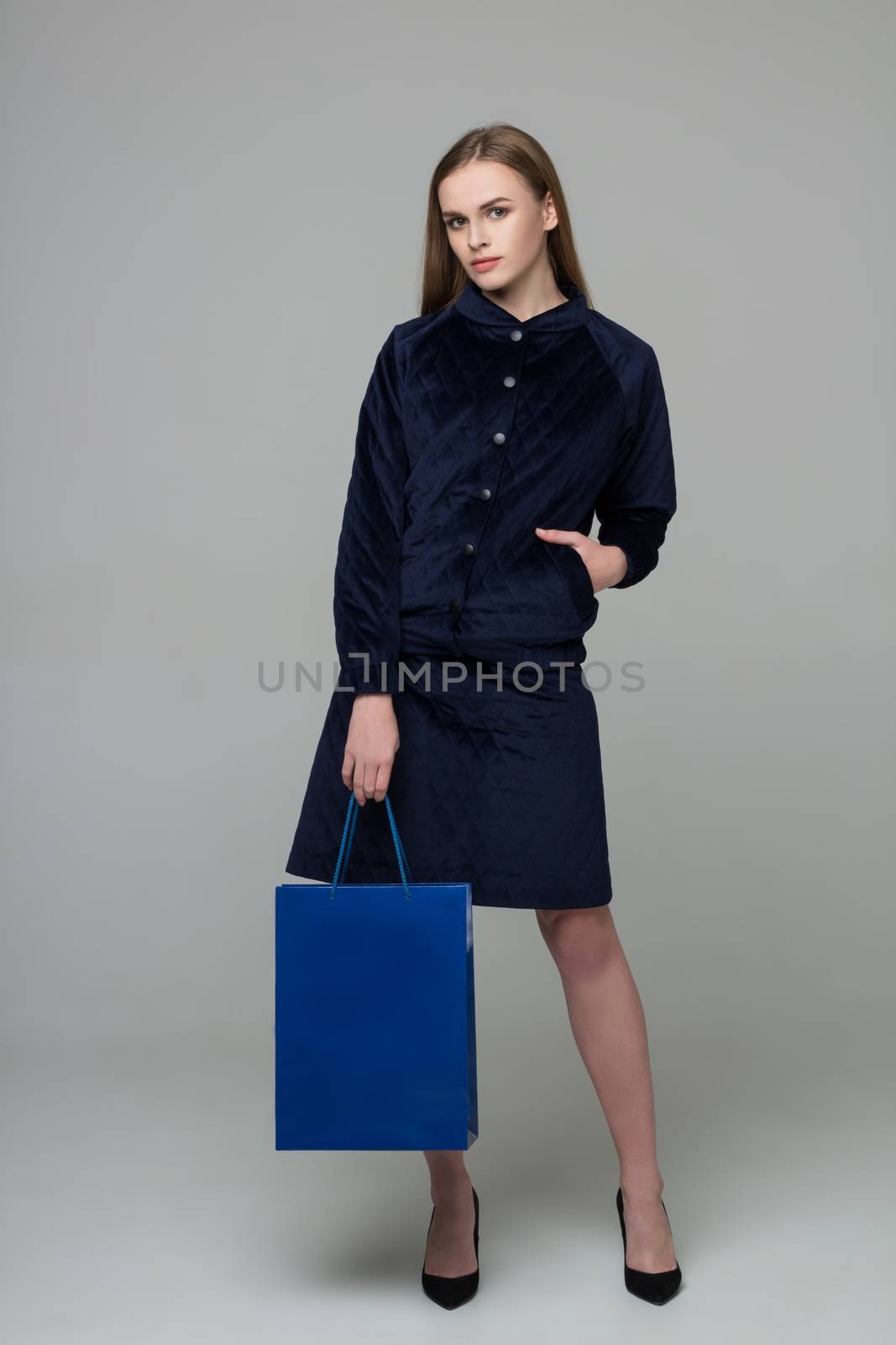 Young model long-haired blond girl in dark skirt suit stands holding blue shopping package