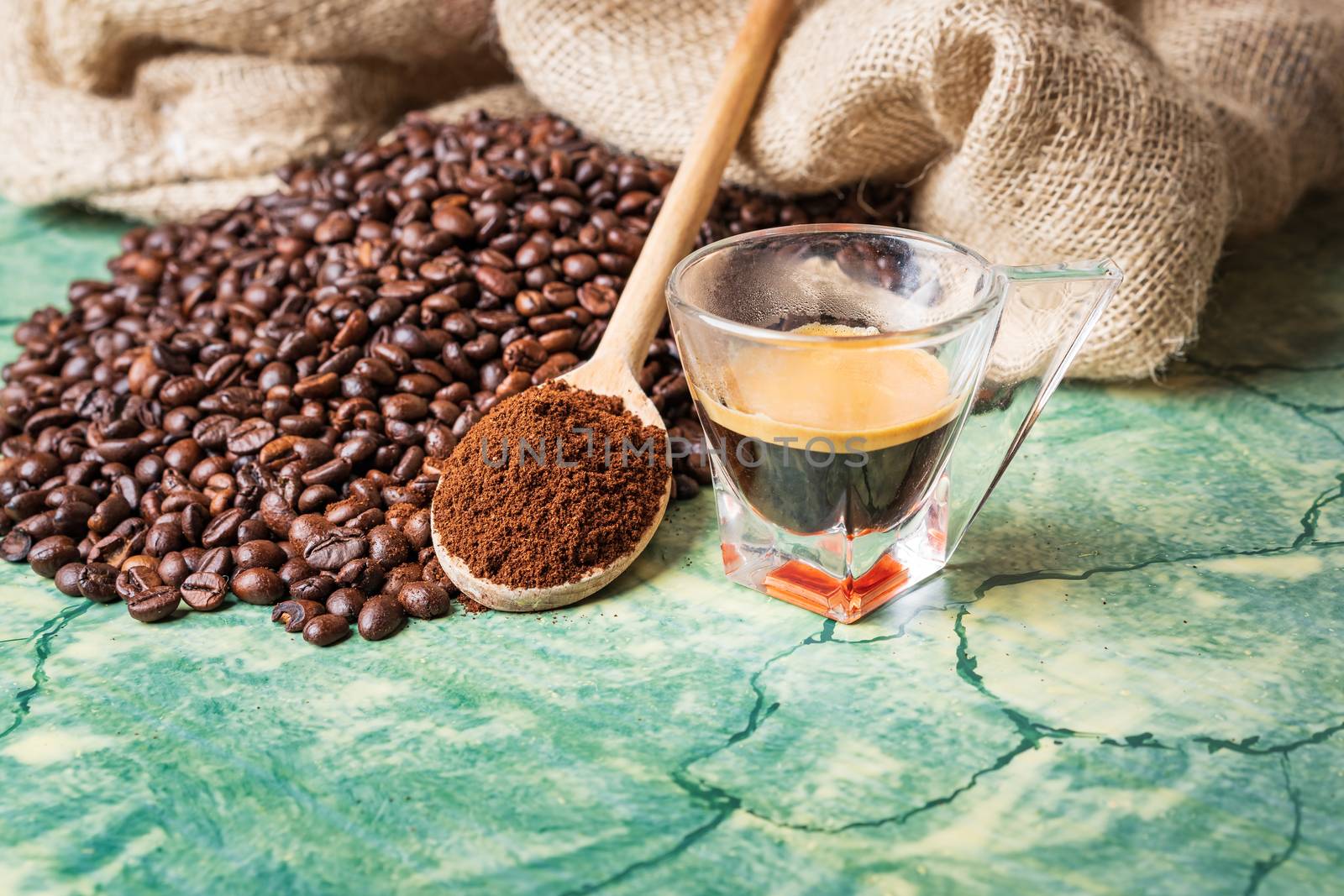 Coffee beans in coffee burlap bag on green table,wooden spoon with ground coffee on top and coffee glass cup.