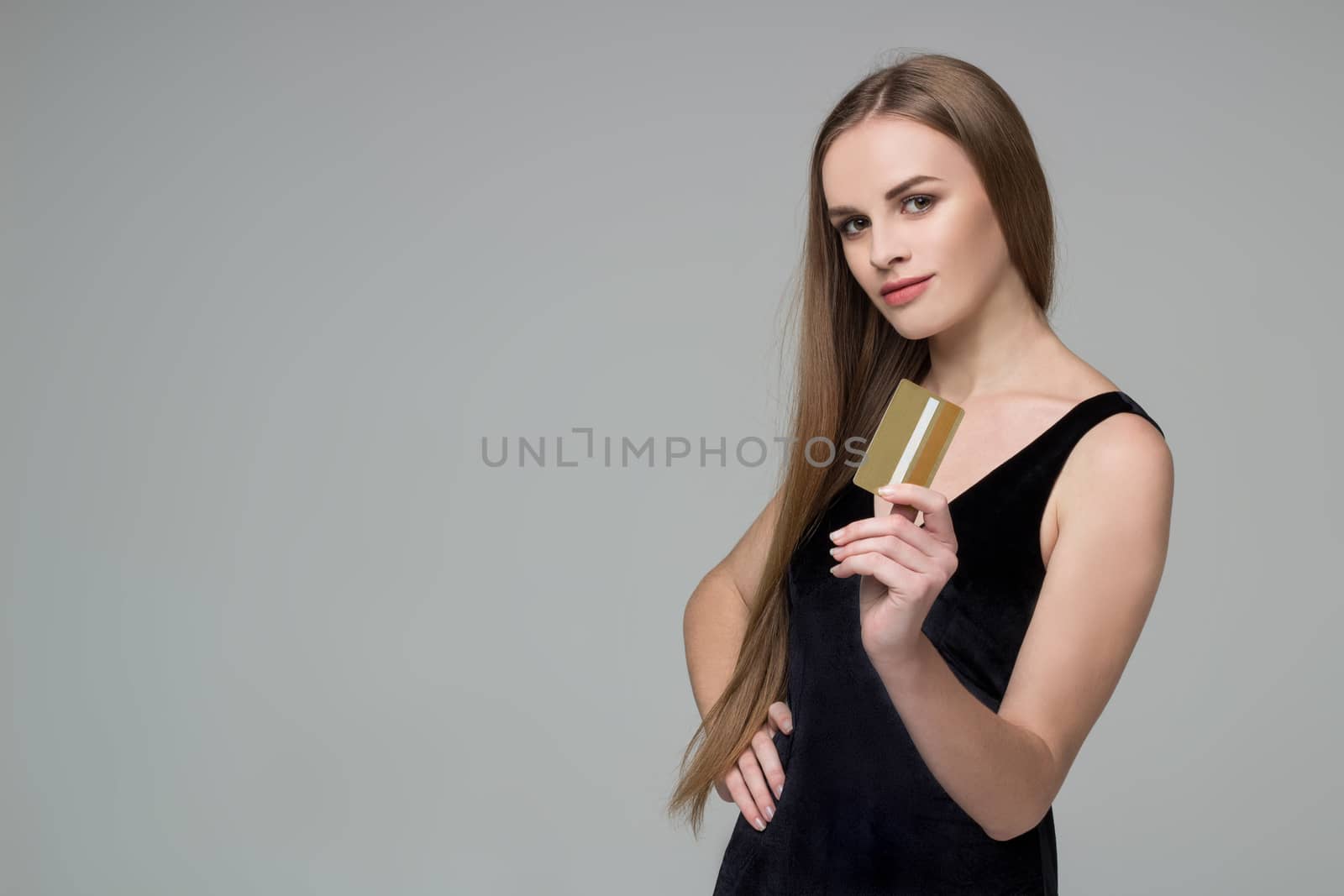 Young model long-haired blond girl in black dress holds golden banking credit card for shopping payment