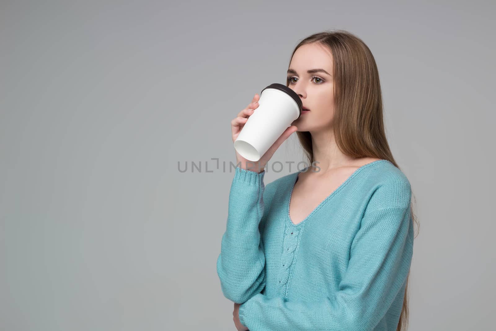 Young model long-haired blond girl in blue aquamarine jamper holds paper plastic coffee glass drinking