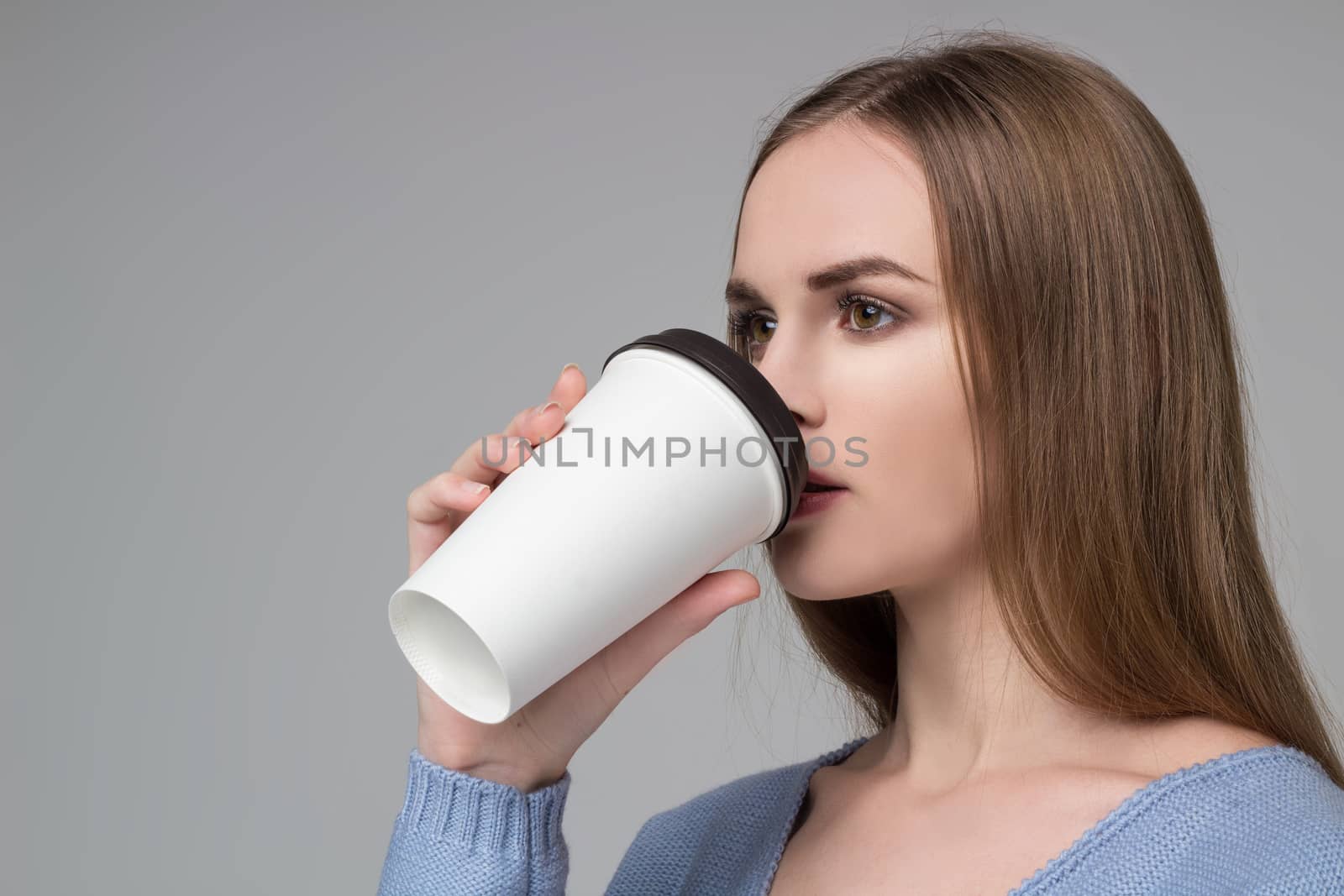 Young model long-haired blond girl in blue purple jamper holds paper plastic coffee glass drinking