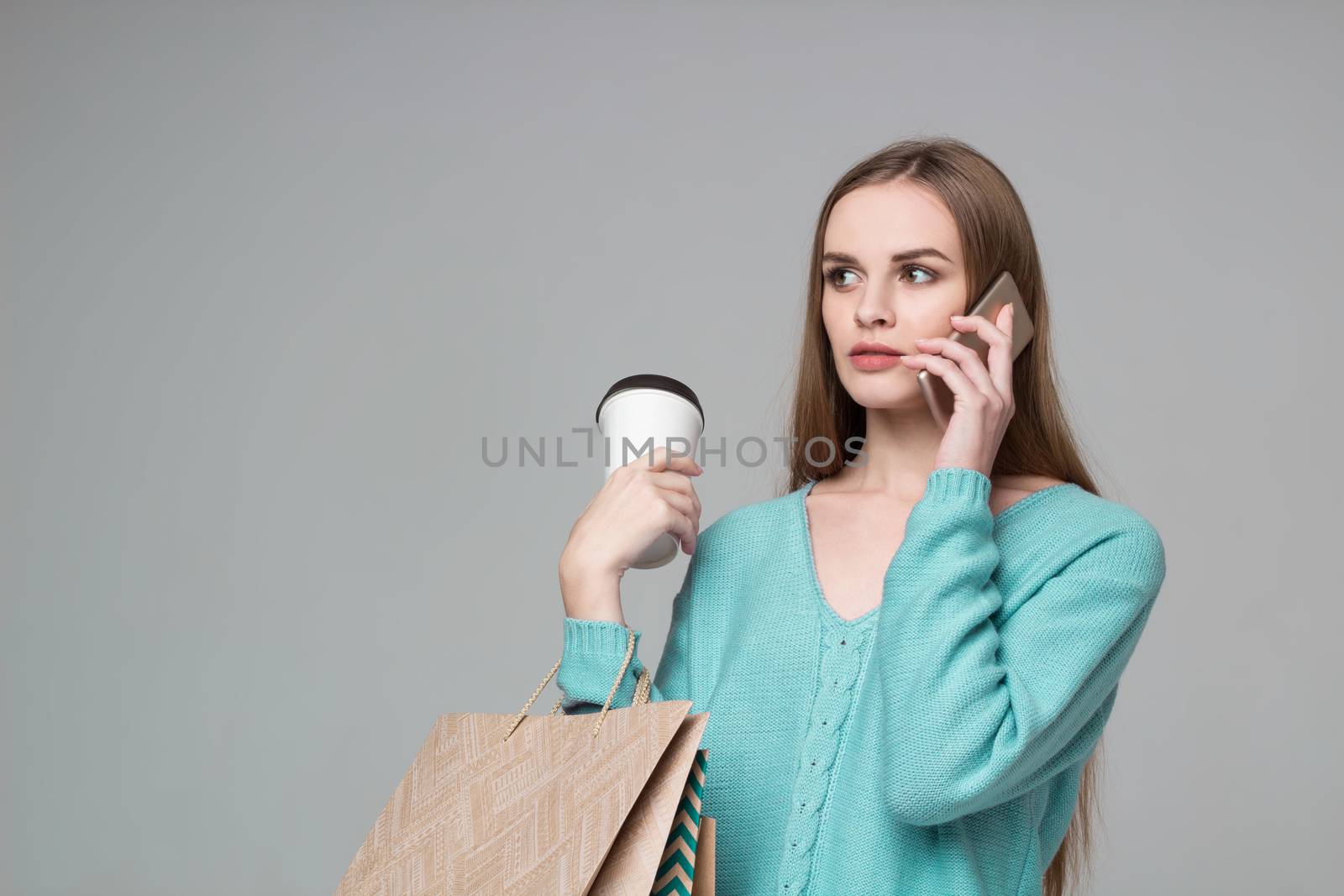 Young model long-haired blond girl in light blue aquamarine jamper holds shopping paper bags and  plastic coffee glass drinking and talking by moble smartphone