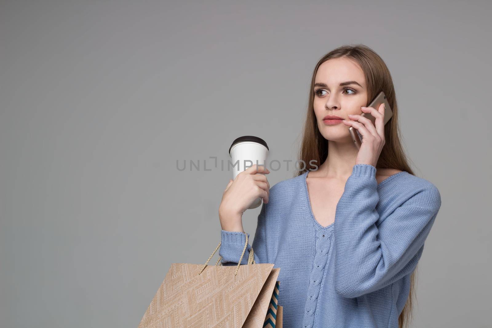 Young model long-haired blond girl in light blue jamper holds shopping paper bags and  plastic coffee glass drinking and talking by moble smartphone