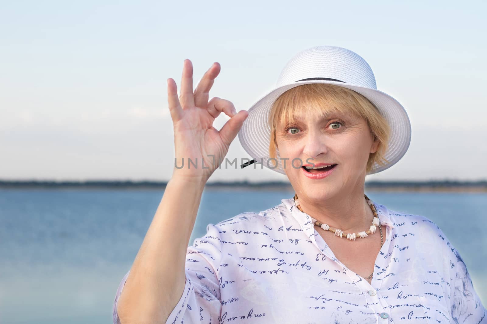Happy smiling retired elderly lady woman in white hat near lake shows okey geste