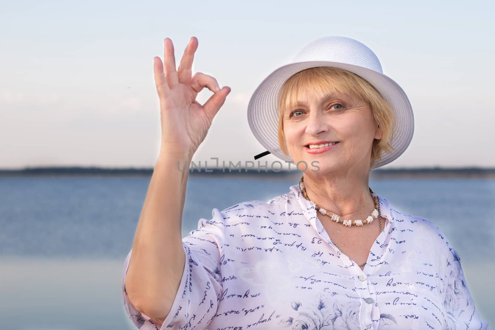 Happy smiling retired elderly lady woman in white hat near lake shows okey geste