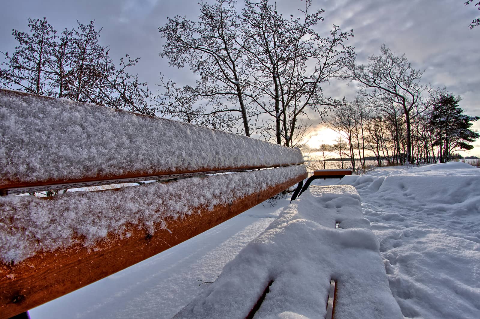 Empty park bench covered in white snow. Beautiful winter day with sun shining through clouds.