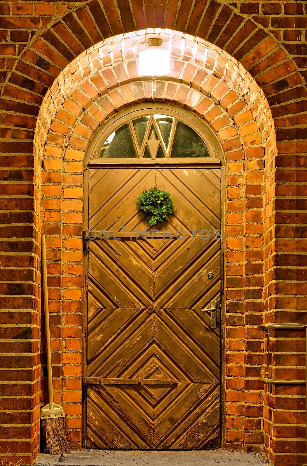 Lighted entrance with old decorated wooden door. Round top and small window above the door. Surrounded with stone walls.