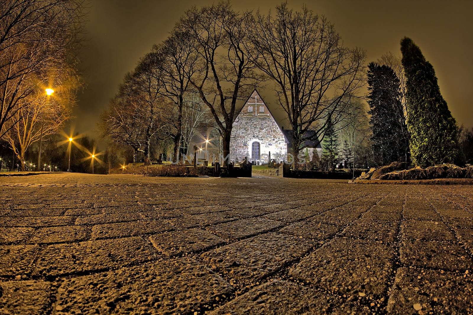 Low and wide angle night photo of a lighted church surrounded with trees. Sidewalk made of stones on the foreground.
