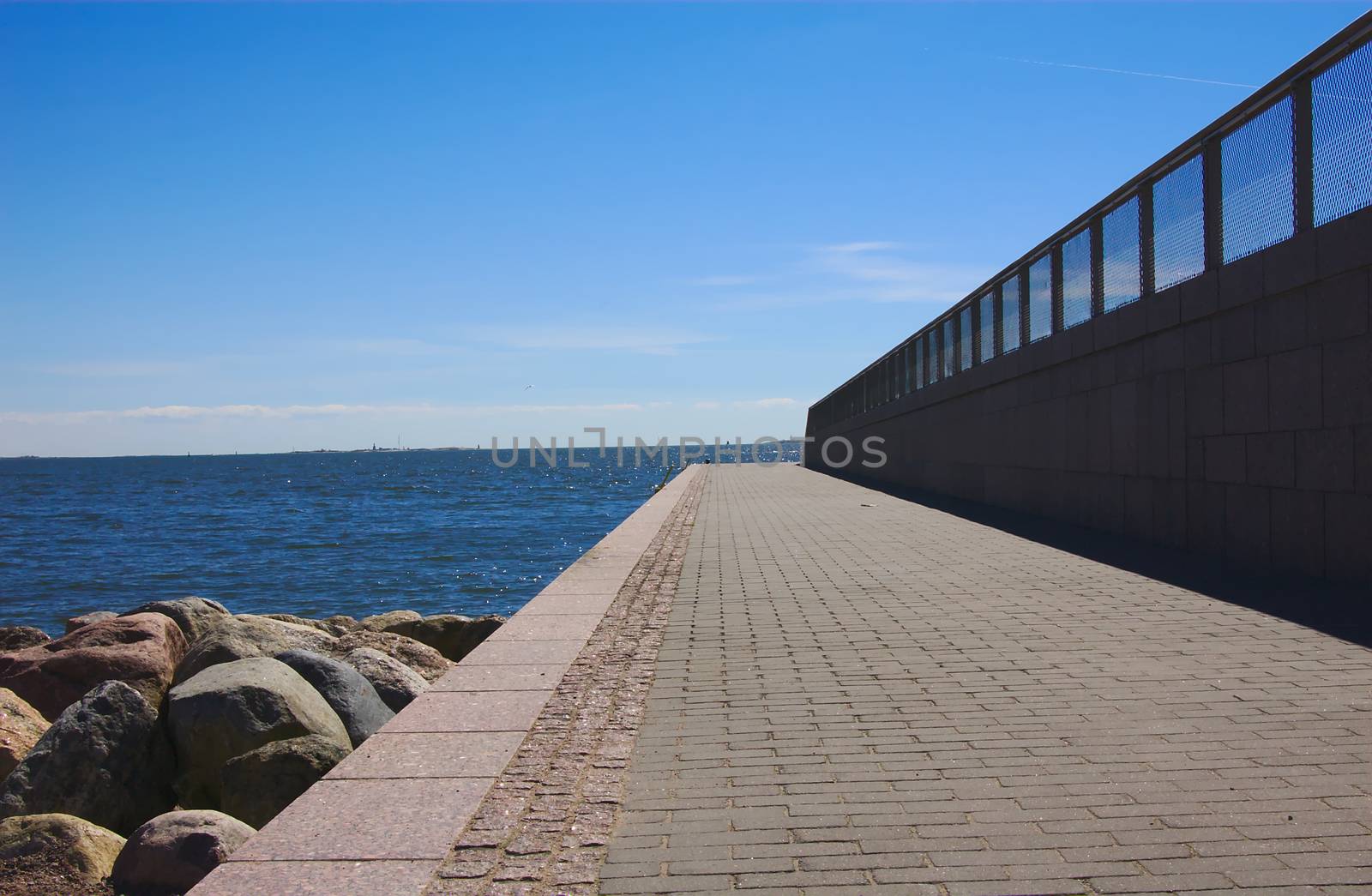 A way to the freedom. Long empty stone sidewalk by the sea at sunny day pointing towards to the horizon. Clean composition with stones, water and blue sky.