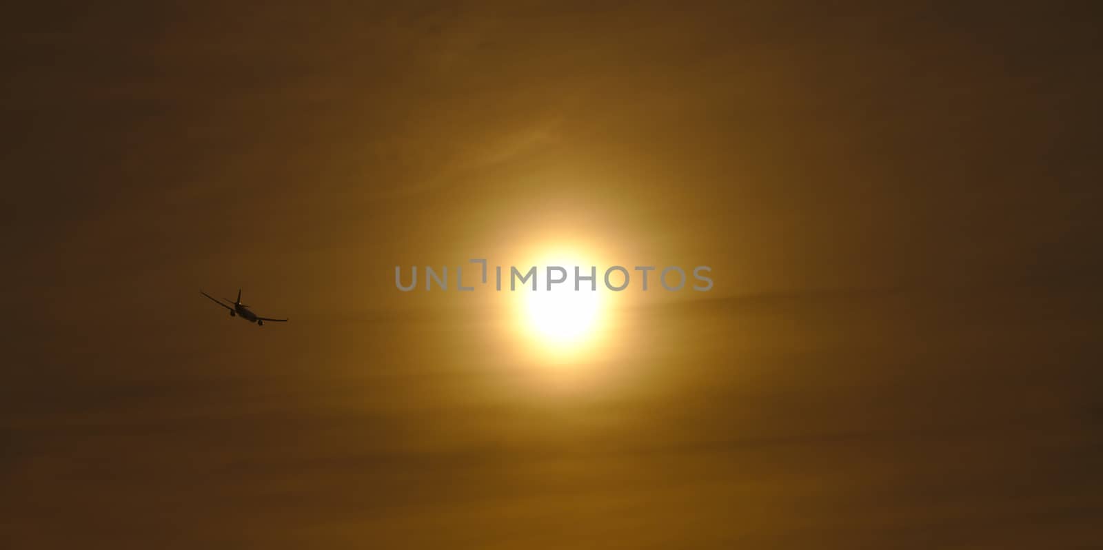 Plane flying away into the evening sun. Dark silhouette of the aircraft visible on the left side of large foggy sun.