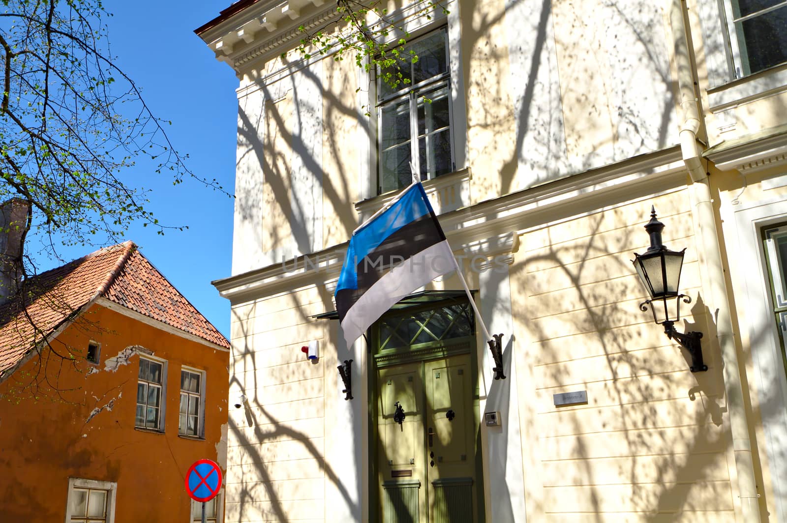 Estonian flag on the wall of a building in the Old Town Tallinn. Beige wall of an old building covered with shadows of a trees. Flag hanging next to the door.