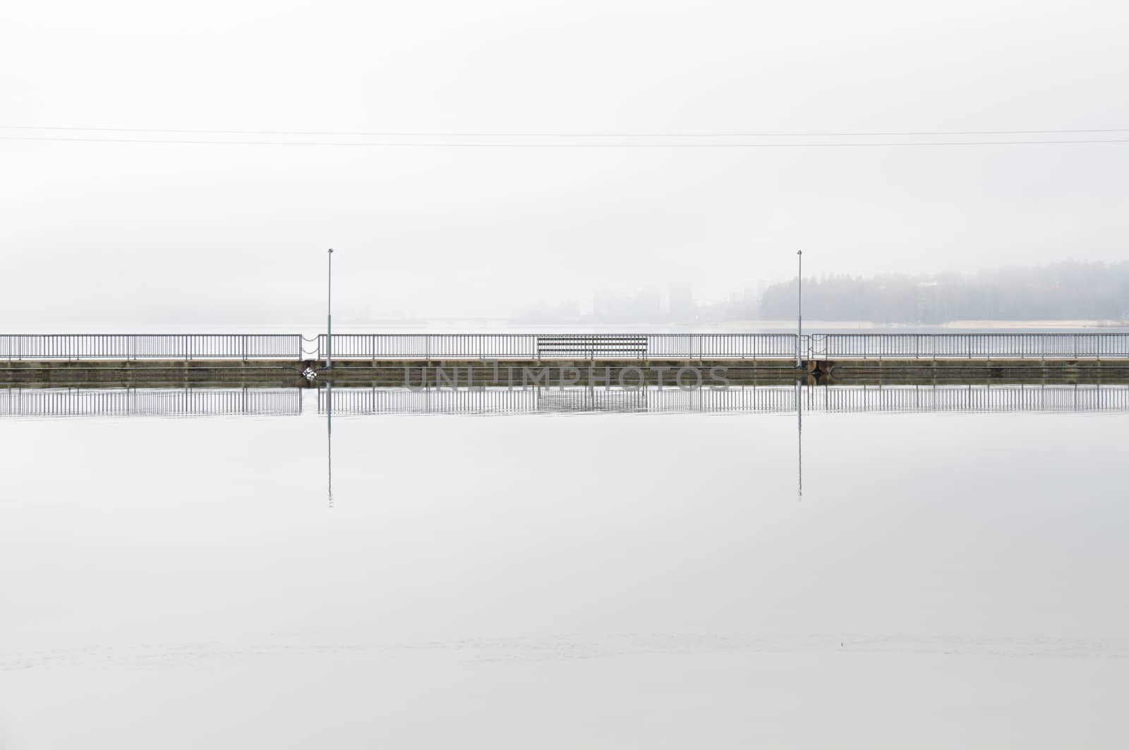 Clear reflection of a walking bridge on the surface of silent water early in the morning. Background covered in fog.