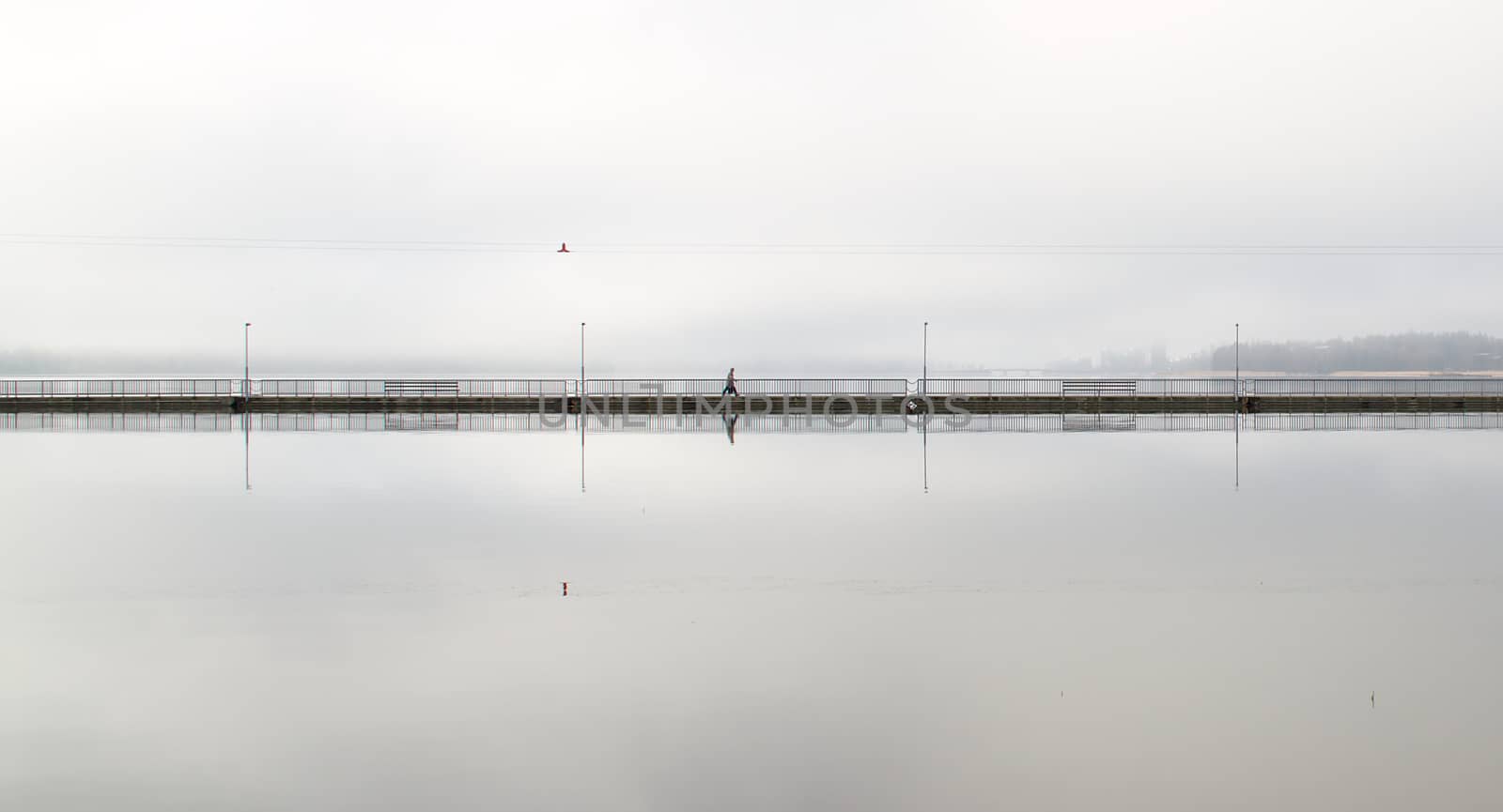 Lonely walker on a long empty bridge early in the morning. Grey and foggy weather in the city.