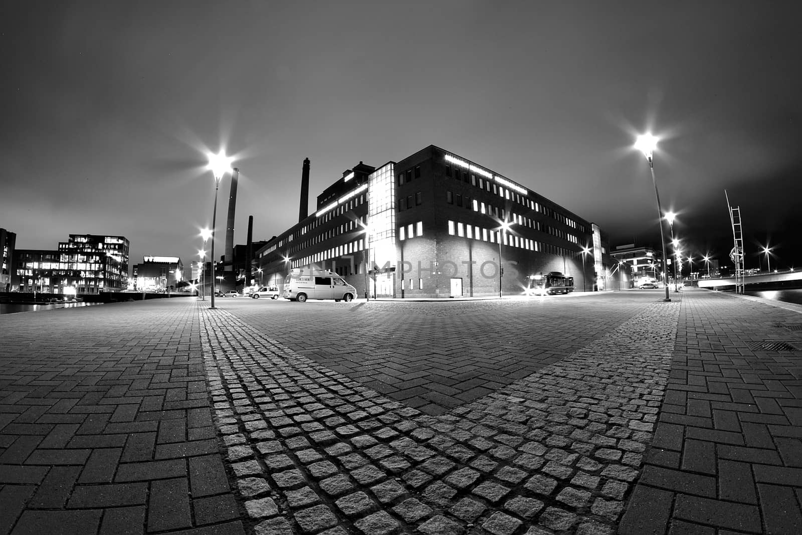 Dramatic low angle black and white night photo of. Large building between lamp posts and stone pavement on foreground.